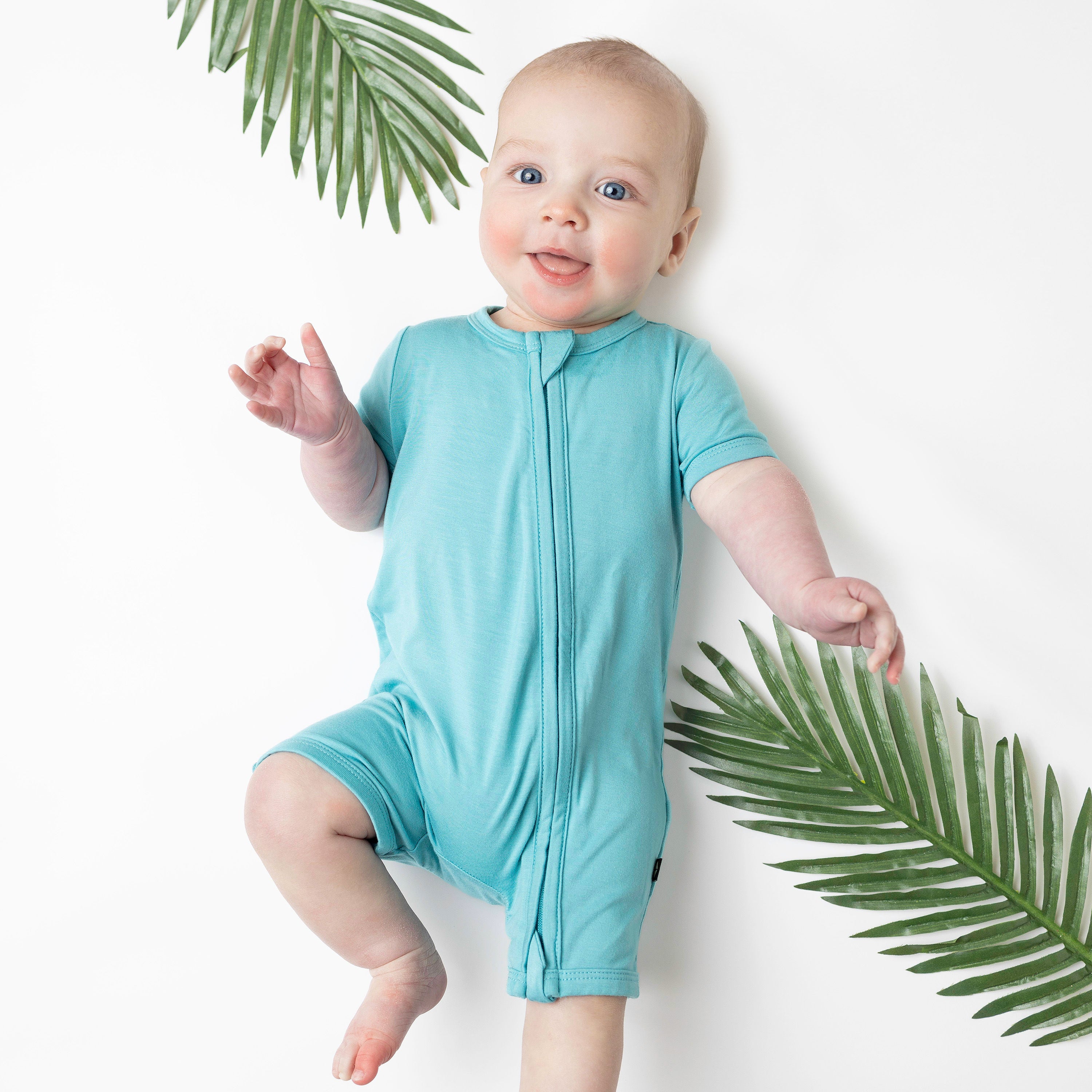 Baby in Zipper Shortall Romper in Makai with palm leaves around