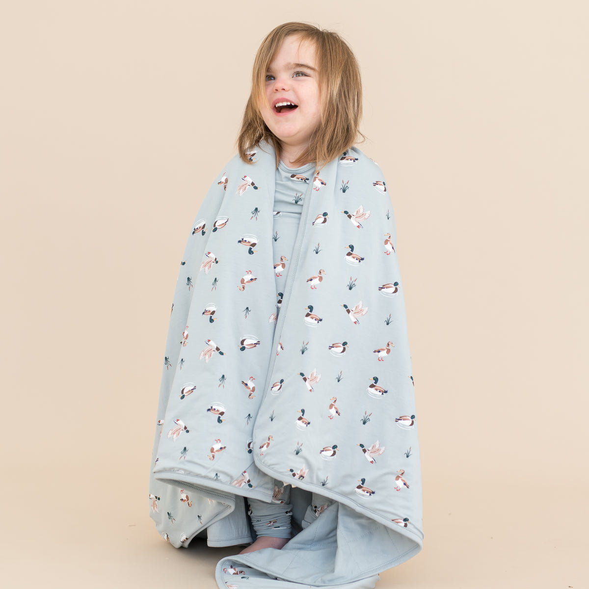 Young child standing in front of a light taupe surface with the Toddler Blanket in Mallard Duck 1.0 wrapped around them
