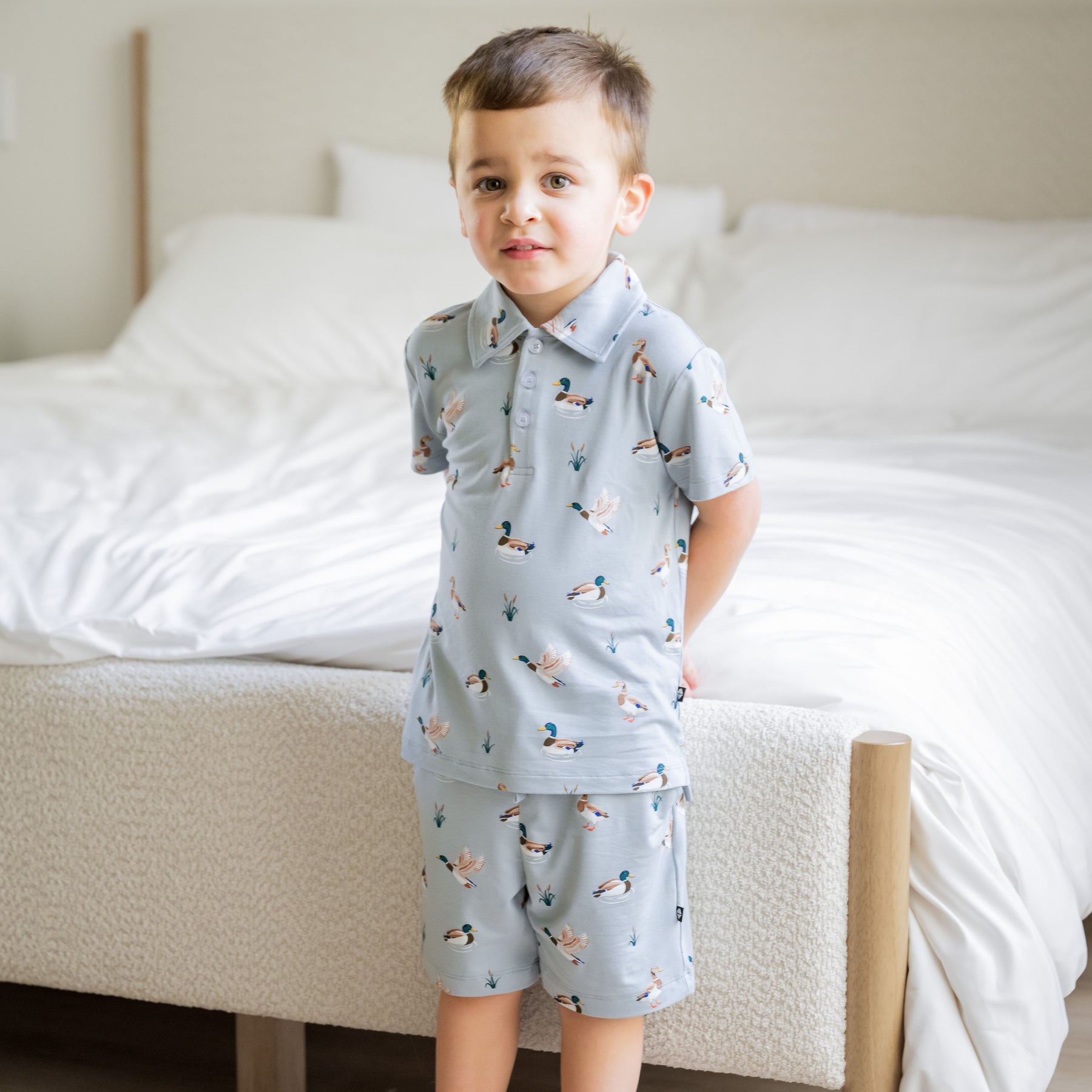 Young boy standing at the edge of a bed wearing the Drawstring Short in Mallard Duck with matching short sleeve polo shirt