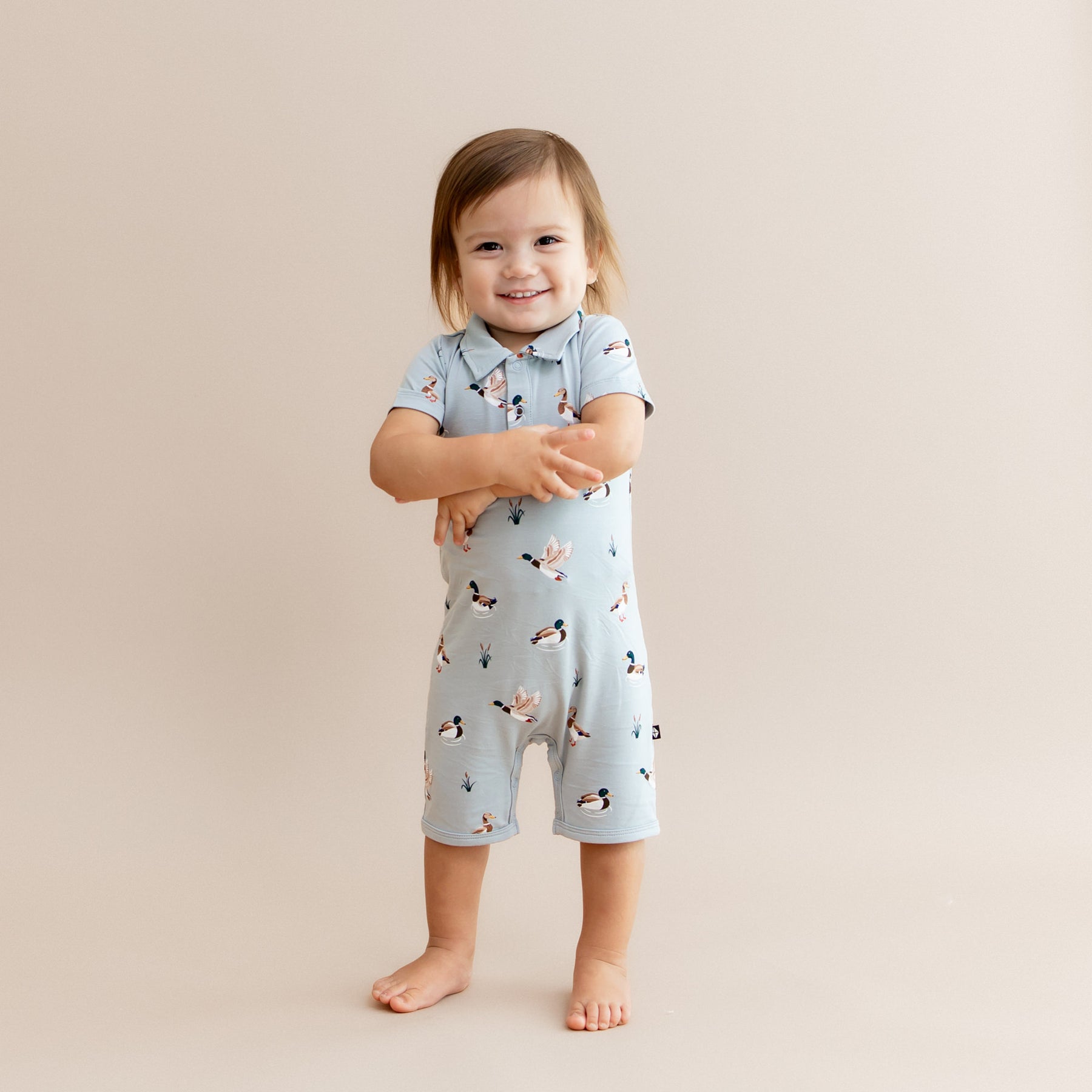 Young smiling toddler standing in front of a light beige background wearing the Polo Shortall in Mallard Duck