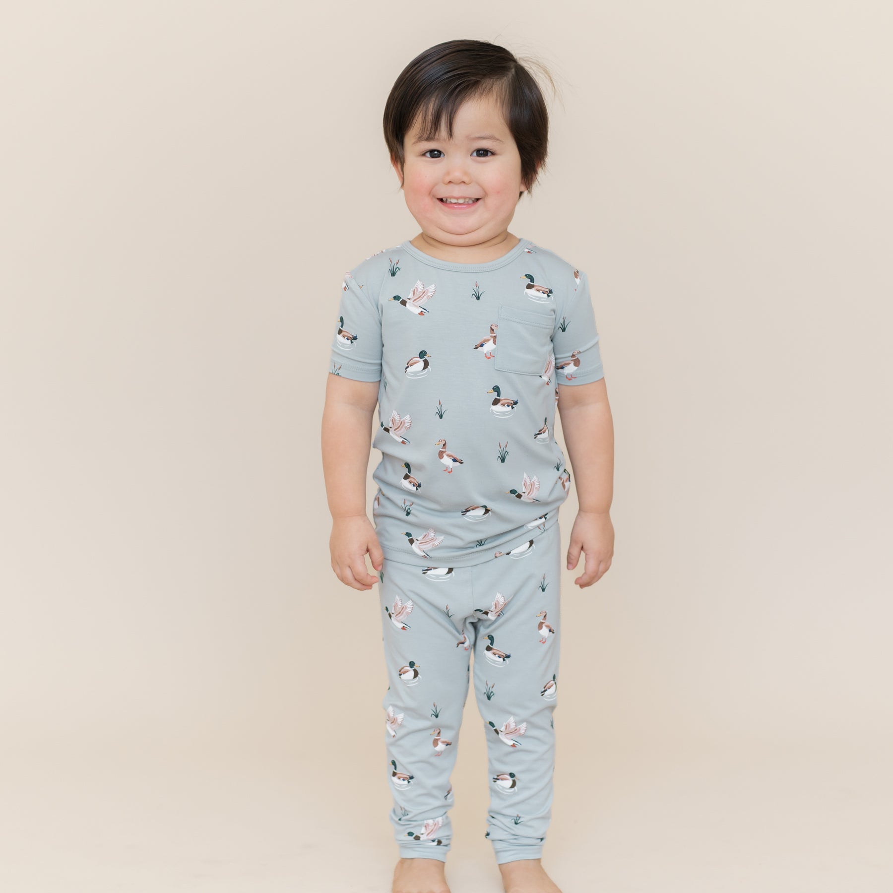 Smiling boy standing in front of a light taupe background wearing the Short Sleeve with Pants Pajamas in Mallard Duck