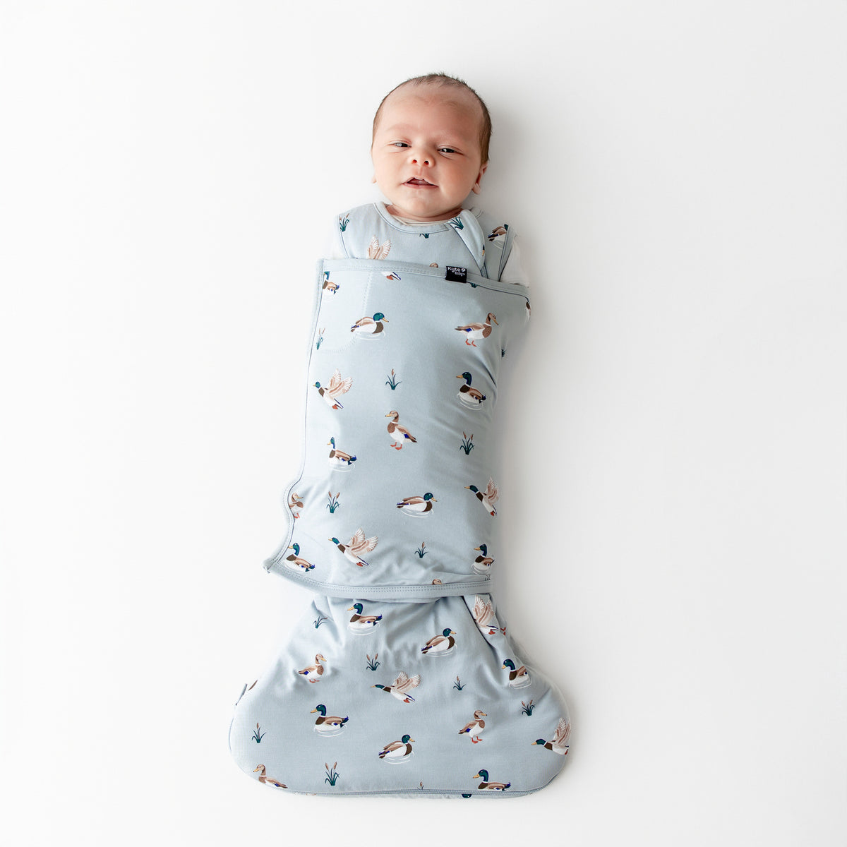 Newborn laying on a white surface swaddled in the Sleep Bag Swaddler in Mallard Duck 1.0