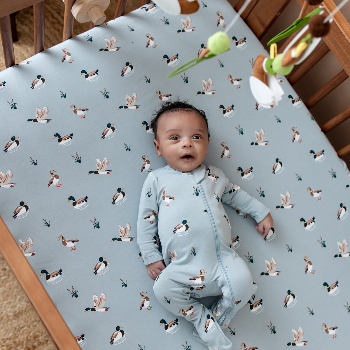 Infant wearing the zippered footie in mallard duck laying in a crib on the Crib Sheet in Mallard Duck