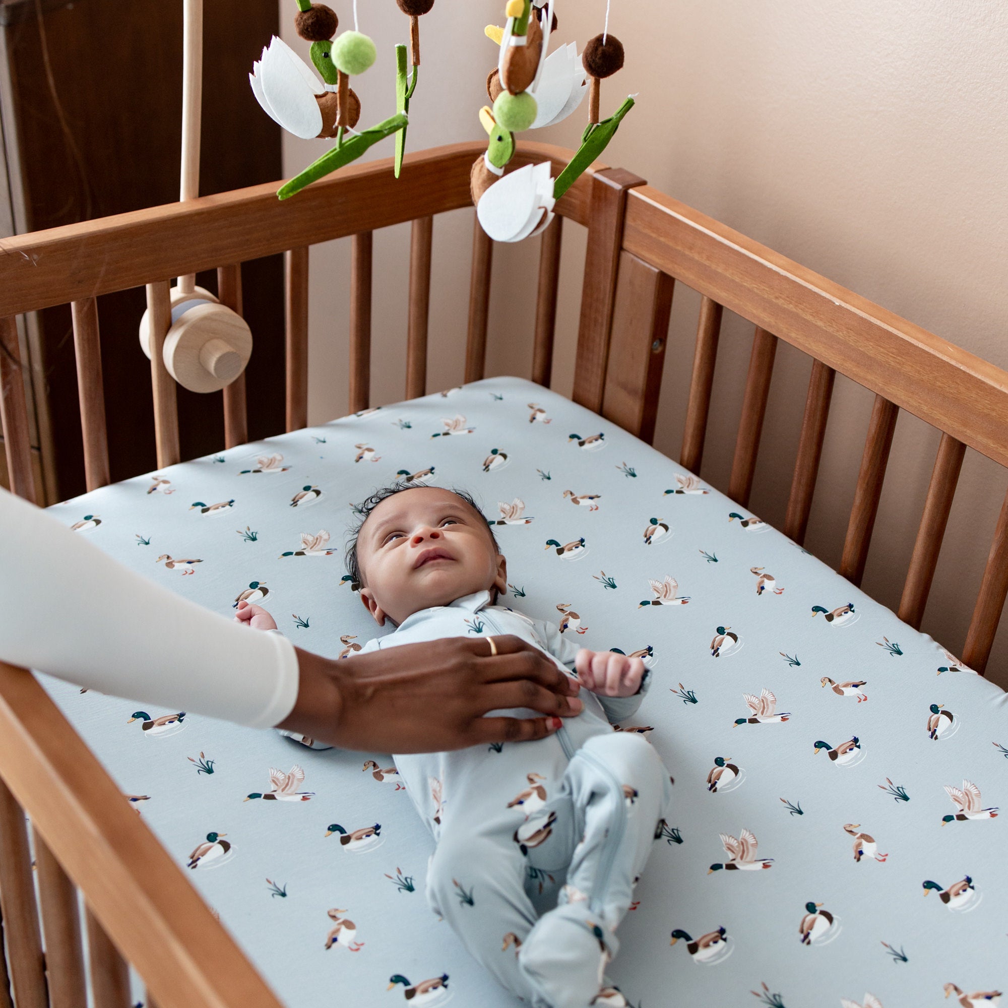 Infant laying in a crib on the Crib Sheet in Mallard Duck looking up at a duck mobile with their mothers hand resting on their stomach