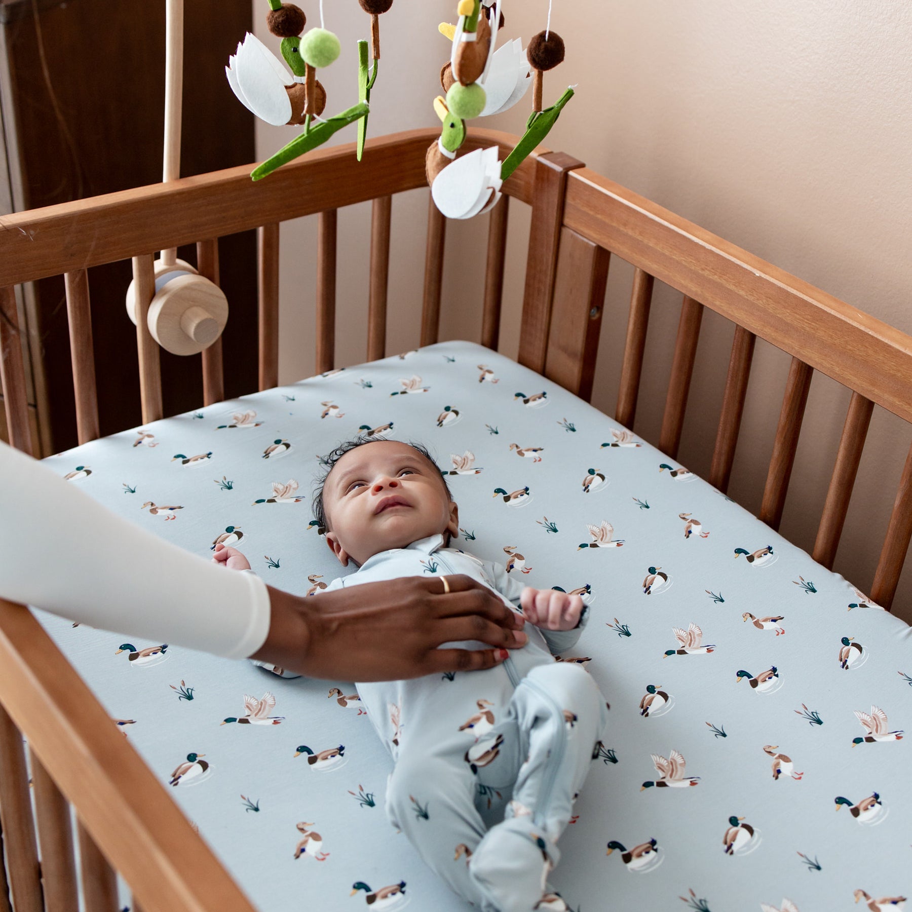 Infant laying in a crib on the Crib Sheet in Mallard Duck looking up at a duck mobile with their mothers hand resting on their stomach