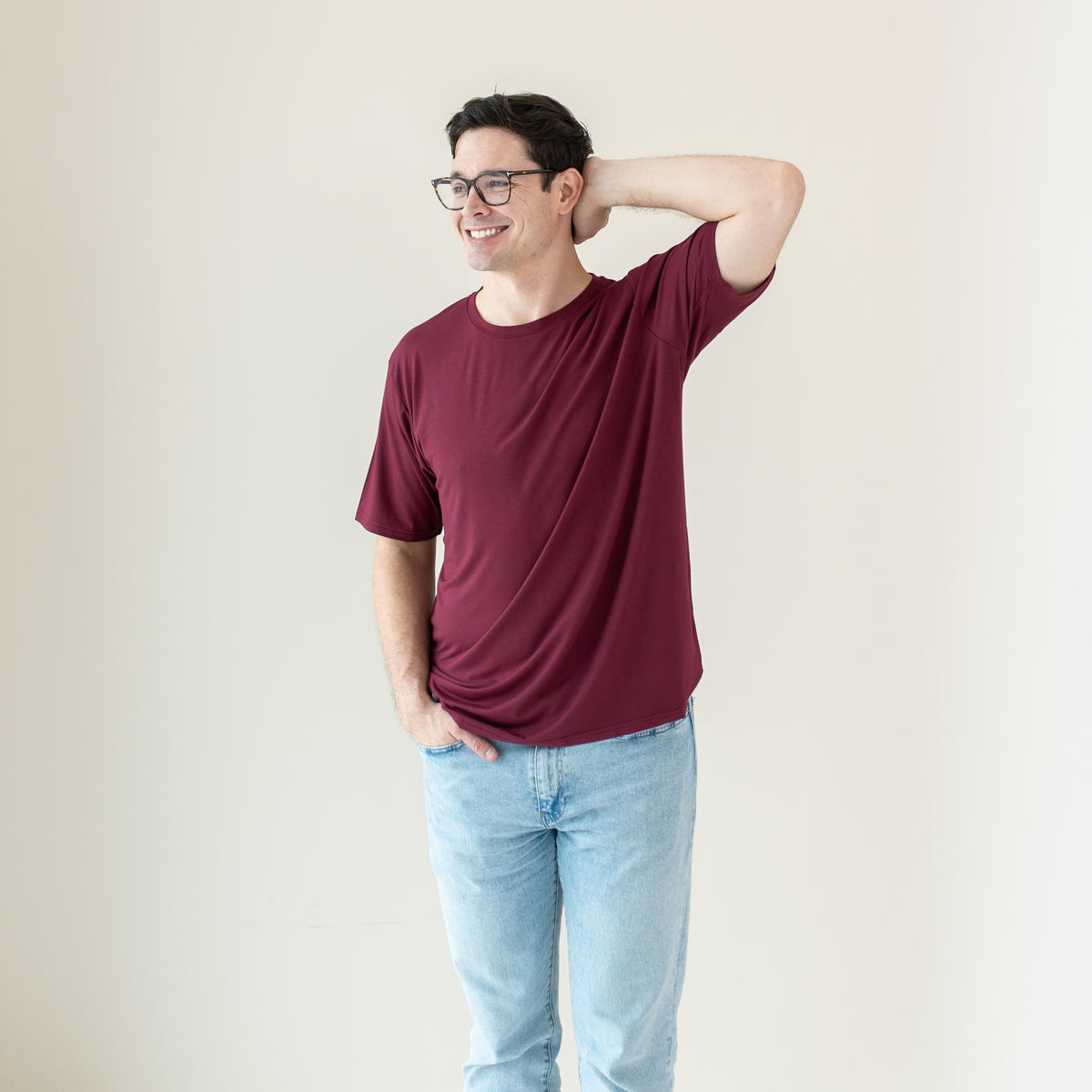 Smiling male model wearing the Men's Crew Neck Tee in Burgundy with one hand behind his head and the other in his jean pocket