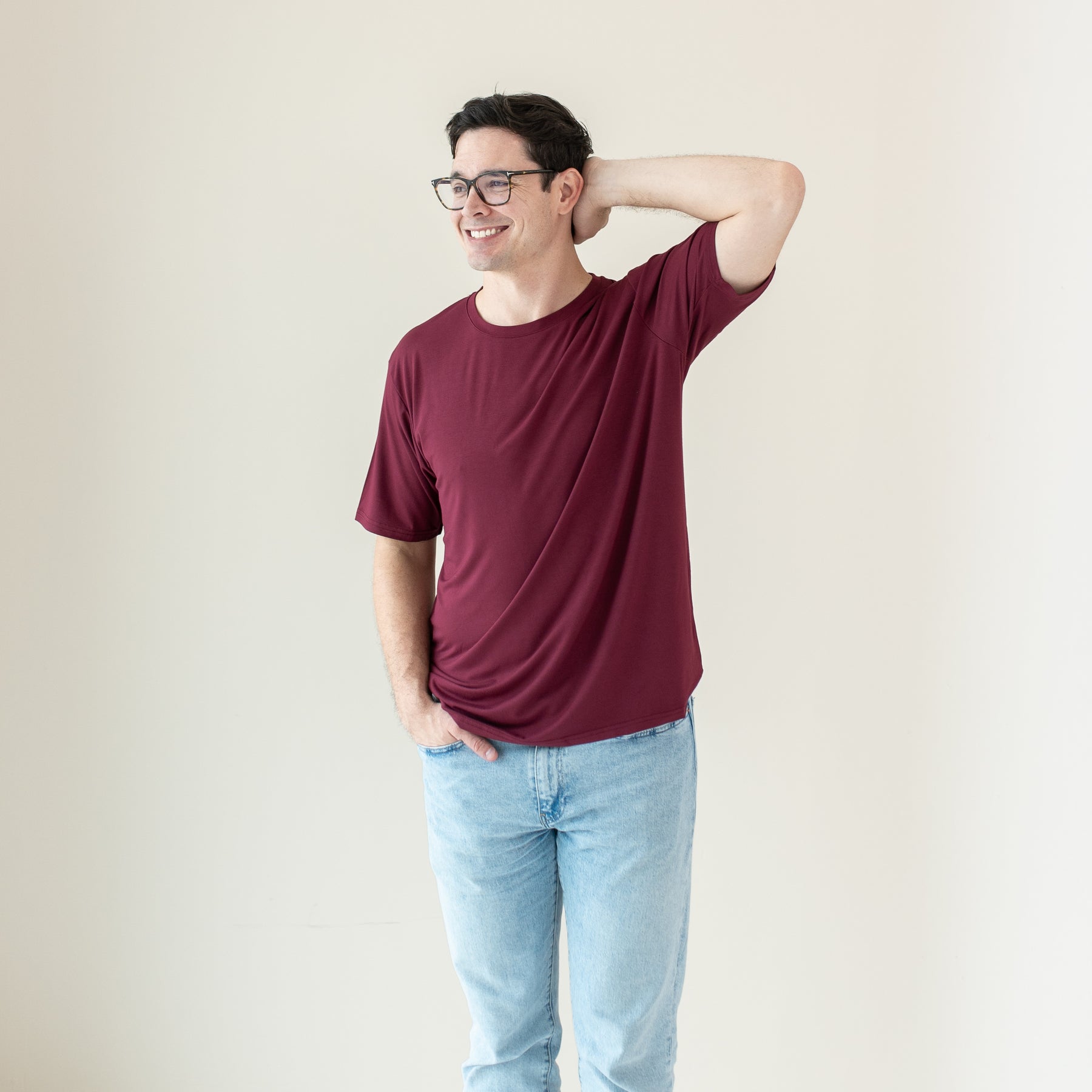 Smiling male model wearing the Men's Crew Neck Tee in Burgundy with one hand behind his head and the other in his jean pocket