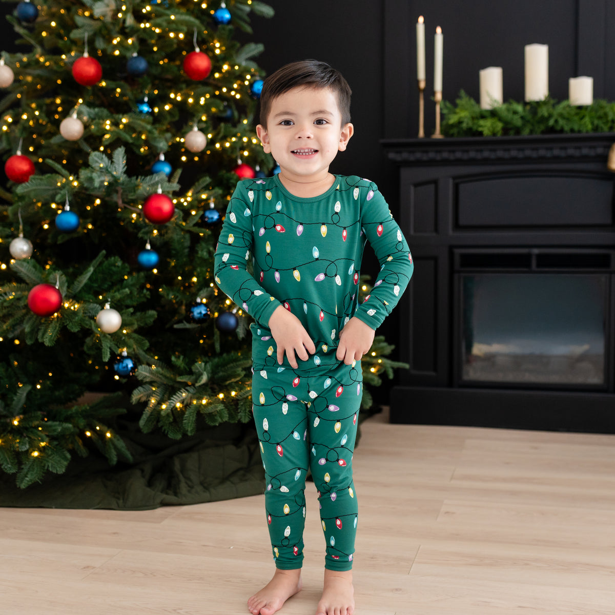 Young boy standing in front of a black fireplace beside a decorated Christmas tree wearing the Long Sleeve Pajamas in Merry and Bright