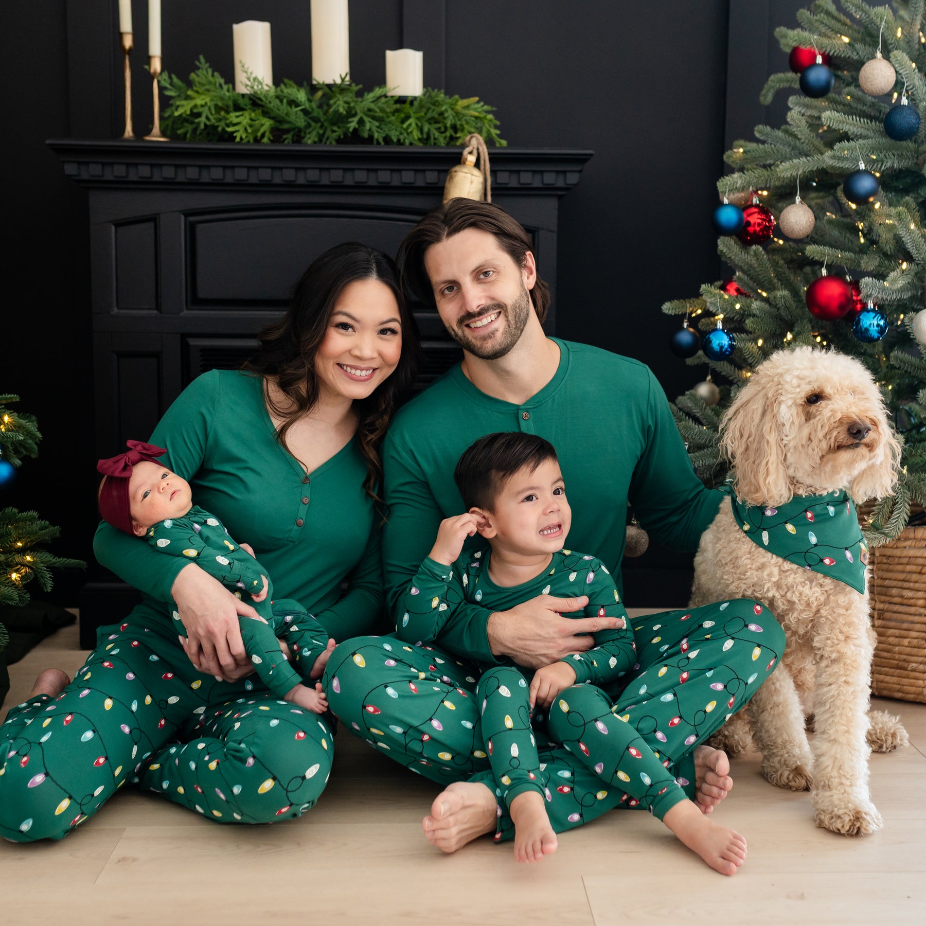 Family of four sitting on the floor matching in various items in the Holiday print Merry and Bright. Medium sized down is sitting beside the family matching in a dog bandana