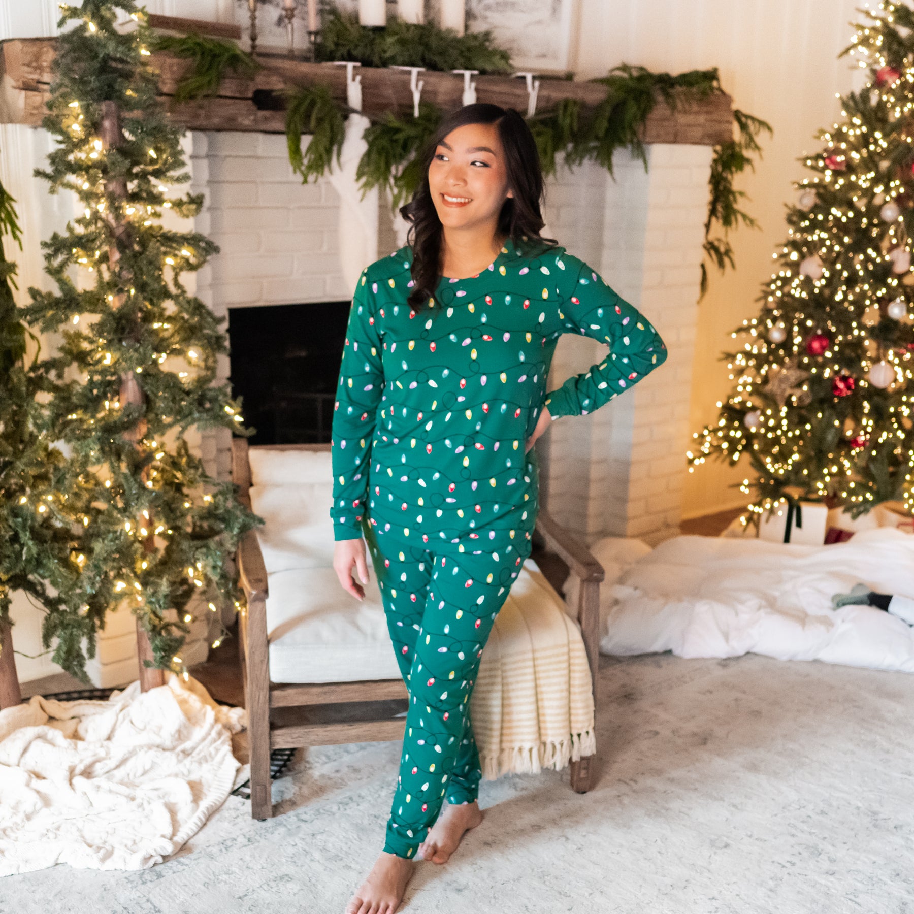 Female model wearing the Women's Jogger Pajama Set in Merry and Bright standing with one hand on hip in front of a white brick fireplace with two lit Christmas trees in the background
