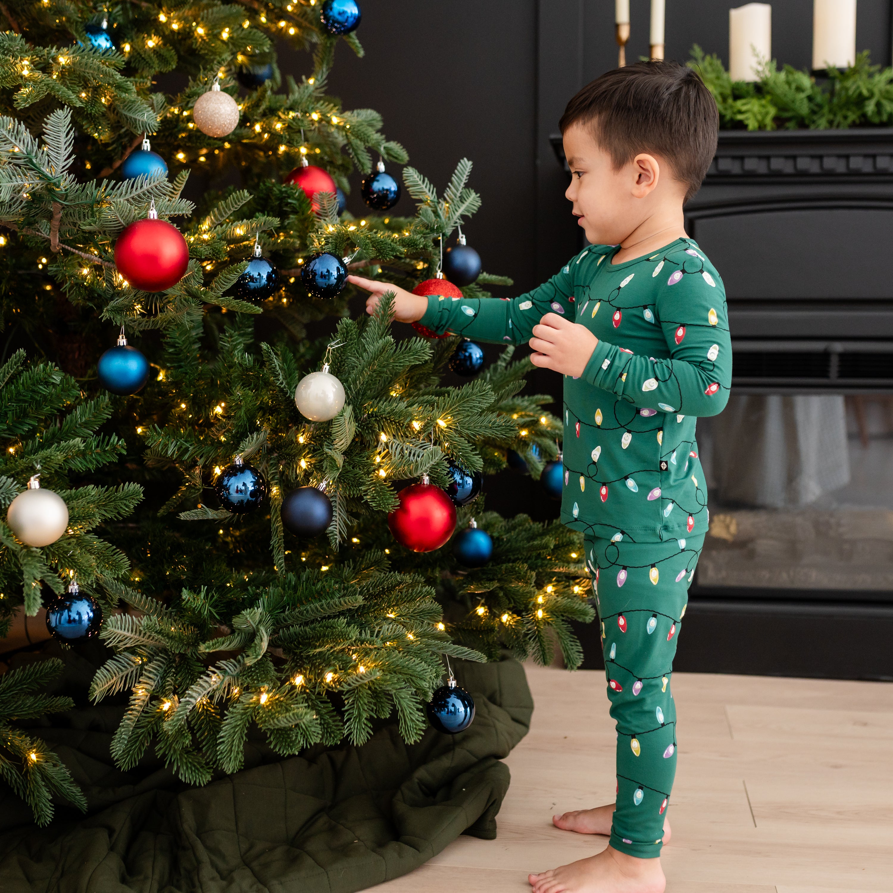 Toddler wearing the Long Sleeve Pajamas in Merry and Bright standing beside a decorated Christmas tree touching an ornament