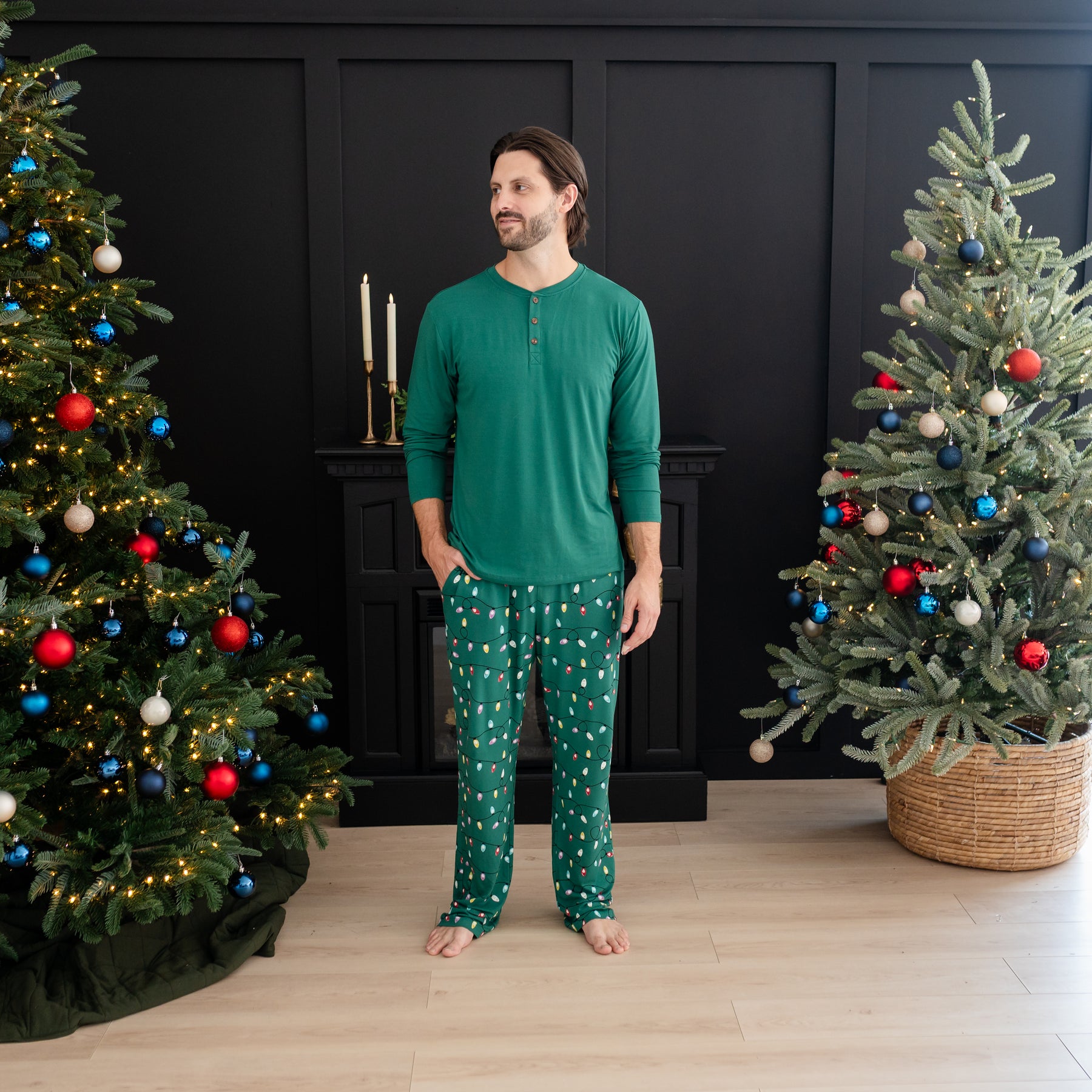 Male model looking off to the side standing with one hand in the pocket of the Men's Lounge Pants in Merry and Bright between two decorated Christmas Trees
