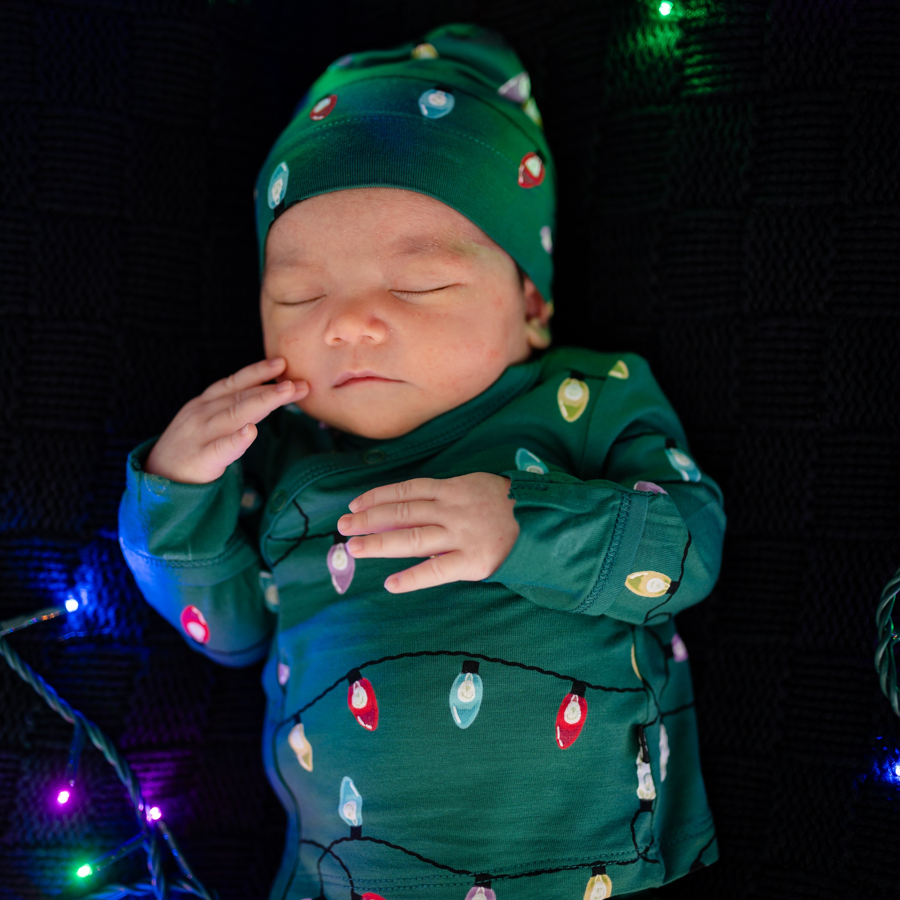 Close up of sleeping newborn wearing the Take Me Home Set with Cap in Merry and Bright surrounded by holiday lights