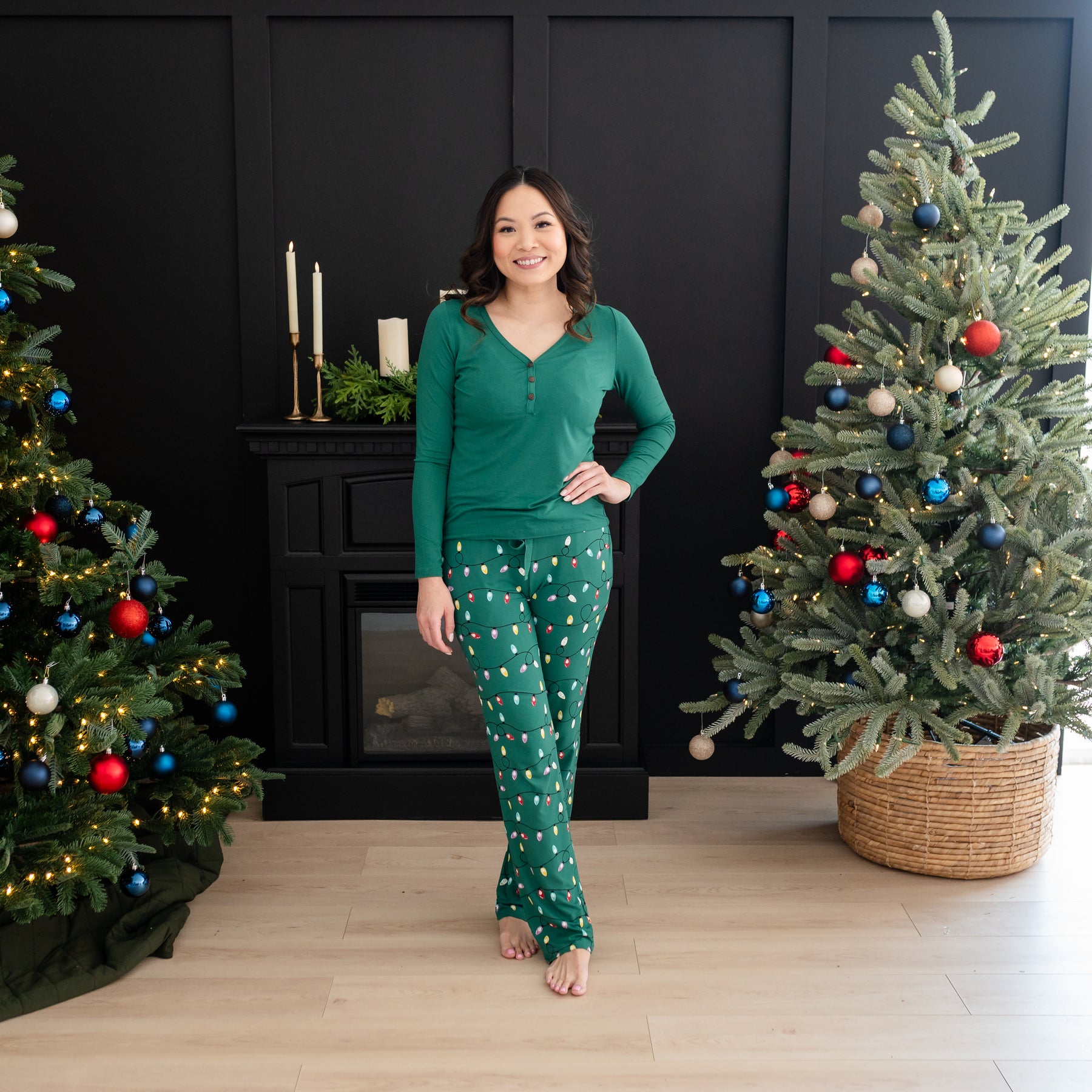 Smiling female standing between two Christmas trees wearing the Women's Lounge Pants in Merry and Bright paired with an Emerald Women's Henley tee