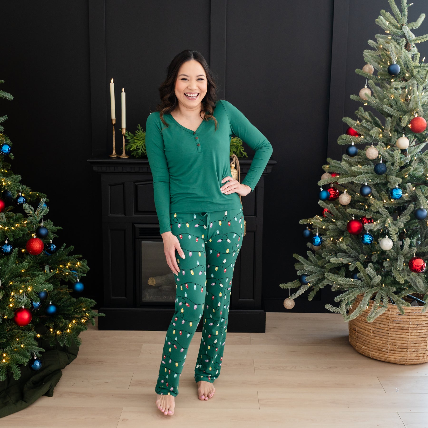 Smiling model standing between two Christmas trees with hand on her hip wearing the Women's Lounge Pants in Merry and Bright and Emerald Long sleeve Henley tee