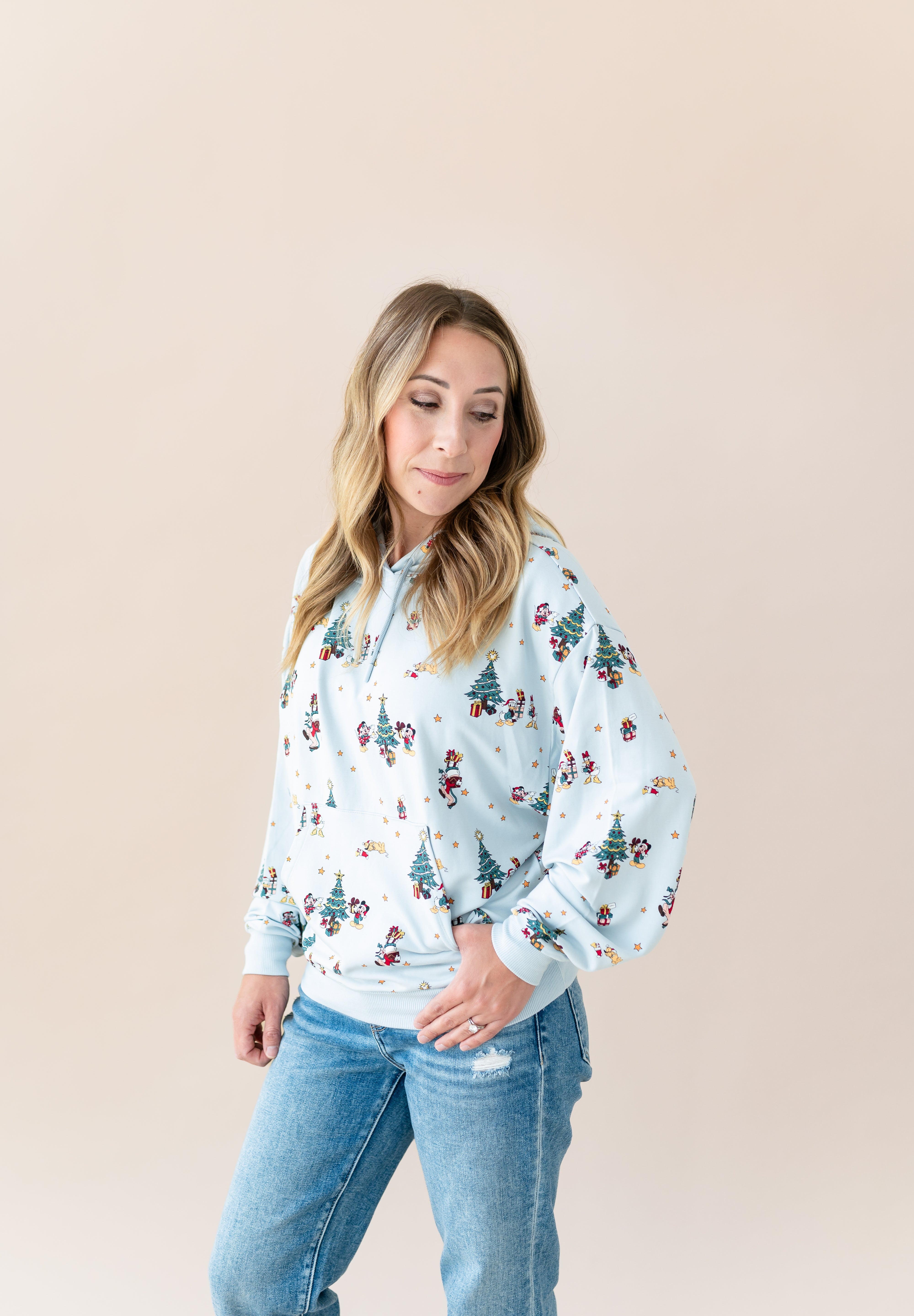 Woman posing in Women's Hoodie in Mickey & Friends Christmas print over blue jeans on a beige background