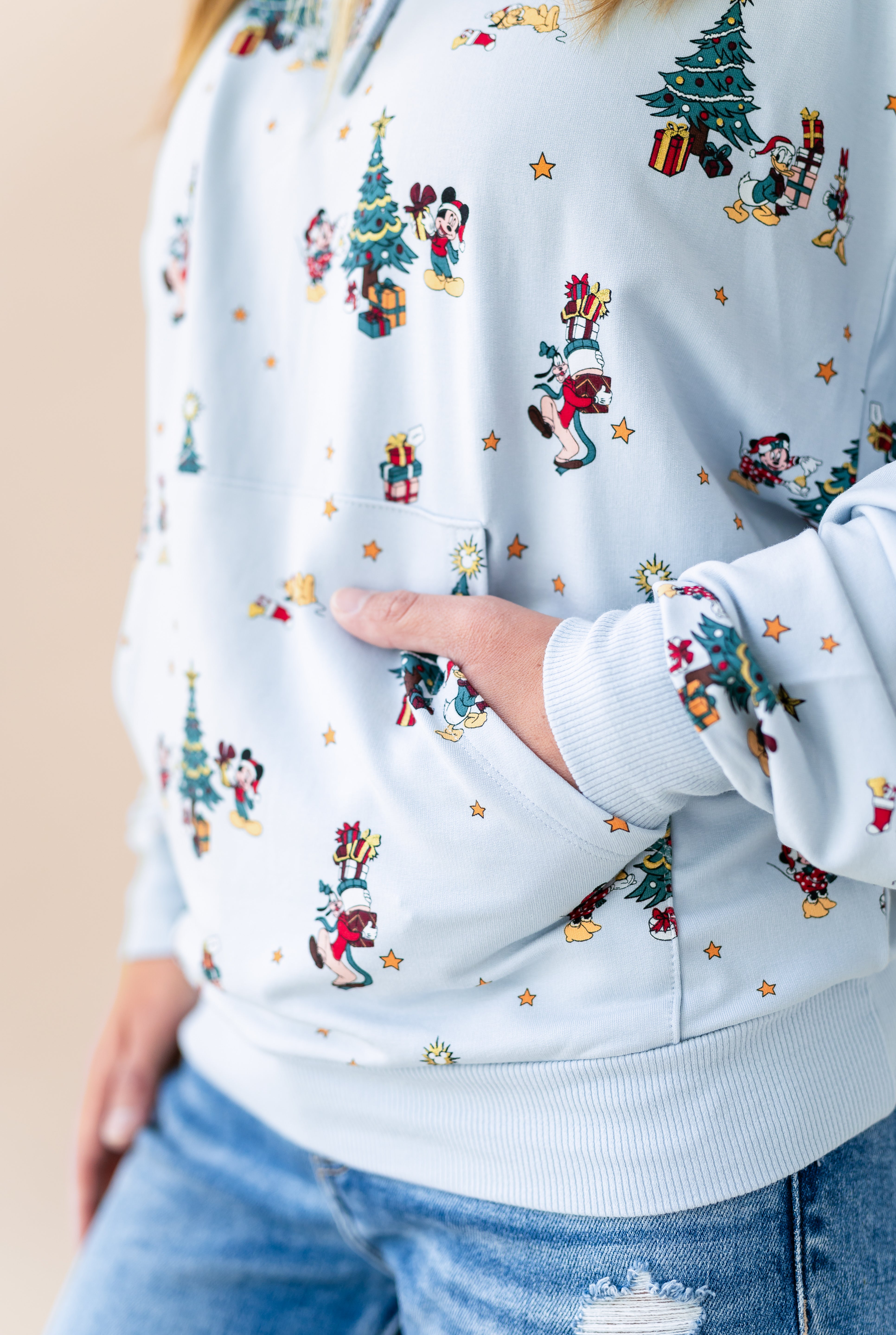 Woman showcasing the pockets on Women's Hoodie in Mickey & Friends Christmas print over blue jeans on a beige background