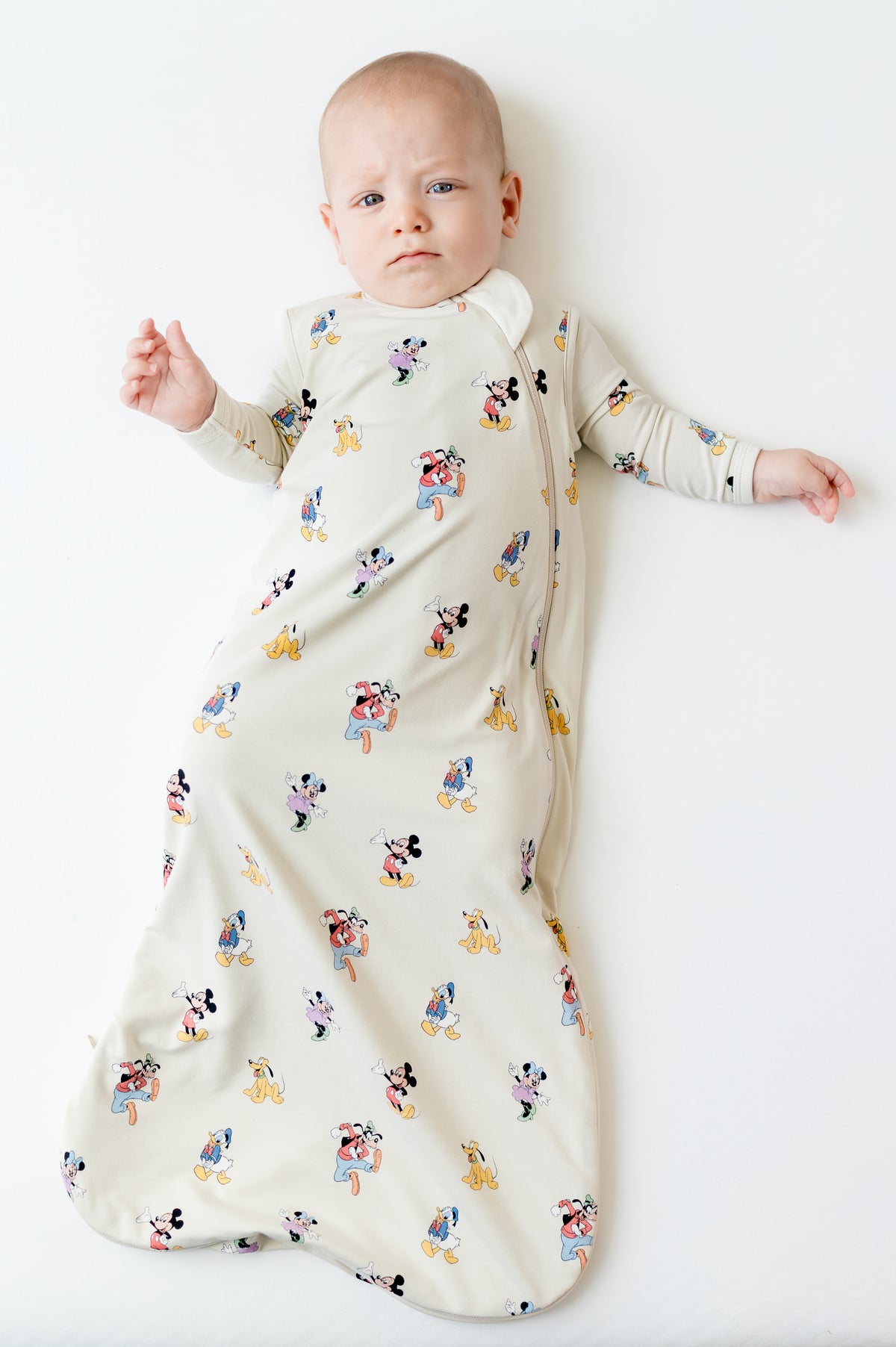 Infant laying on a light neutral surface wearing the Sleep Bag in Mickey Mouse and Friends 0.5 with matching romper