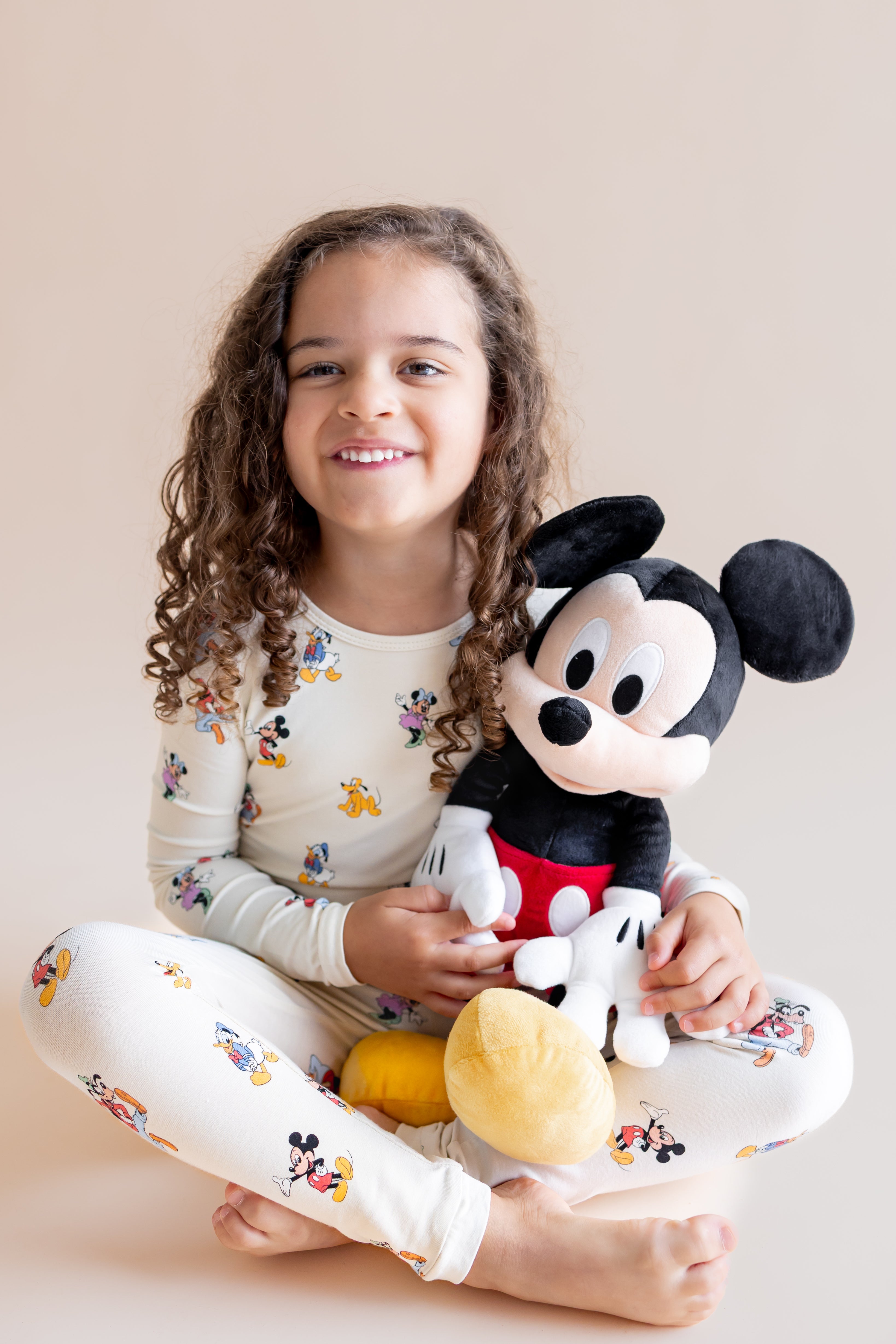 Girl sitting in Long Sleeve Pajamas in Mickey Mouse & Friends print holding a stuff toy on a cream background