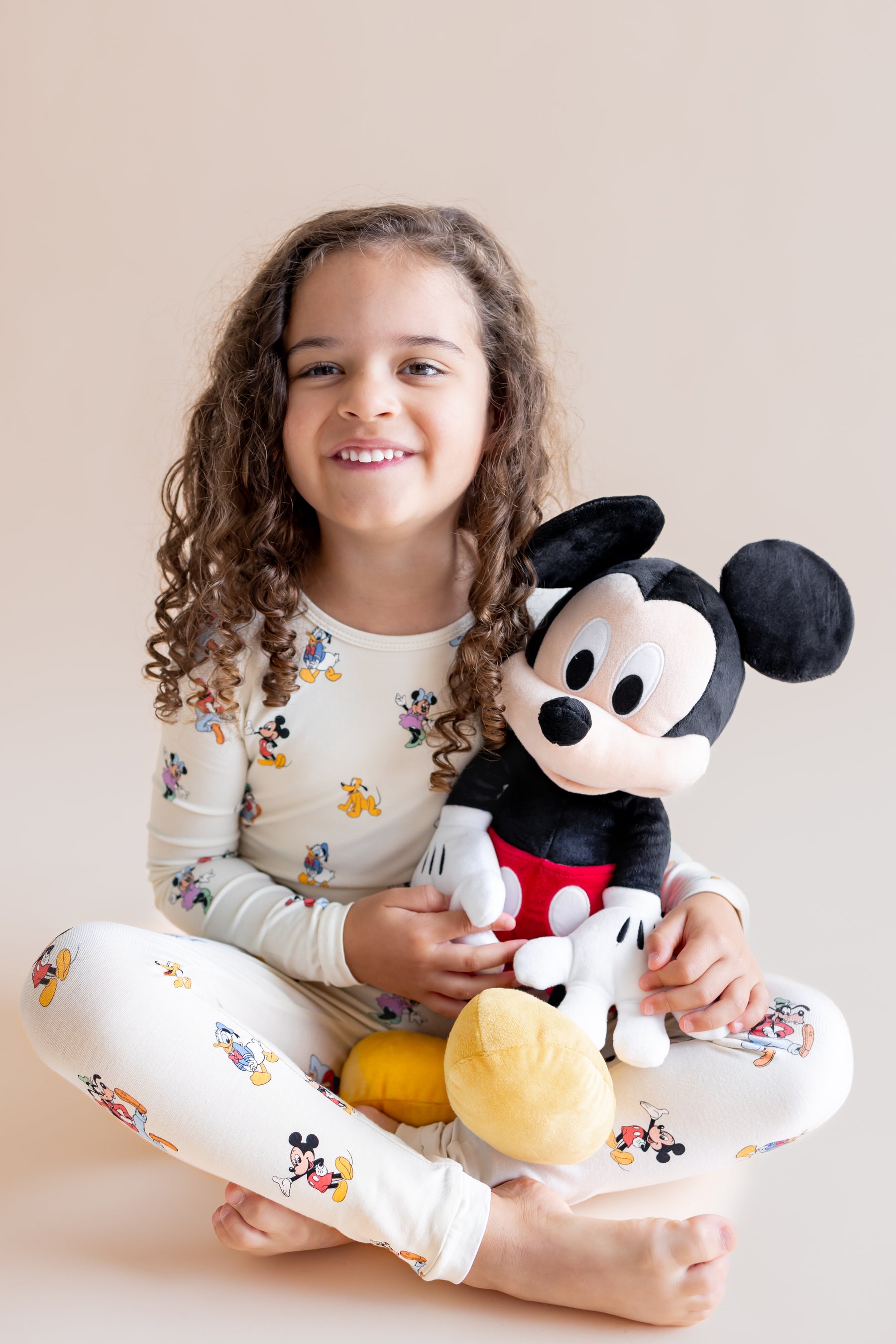 Girl sitting in Long Sleeve Pajamas in Mickey Mouse & Friends print holding a stuff toy on a cream background