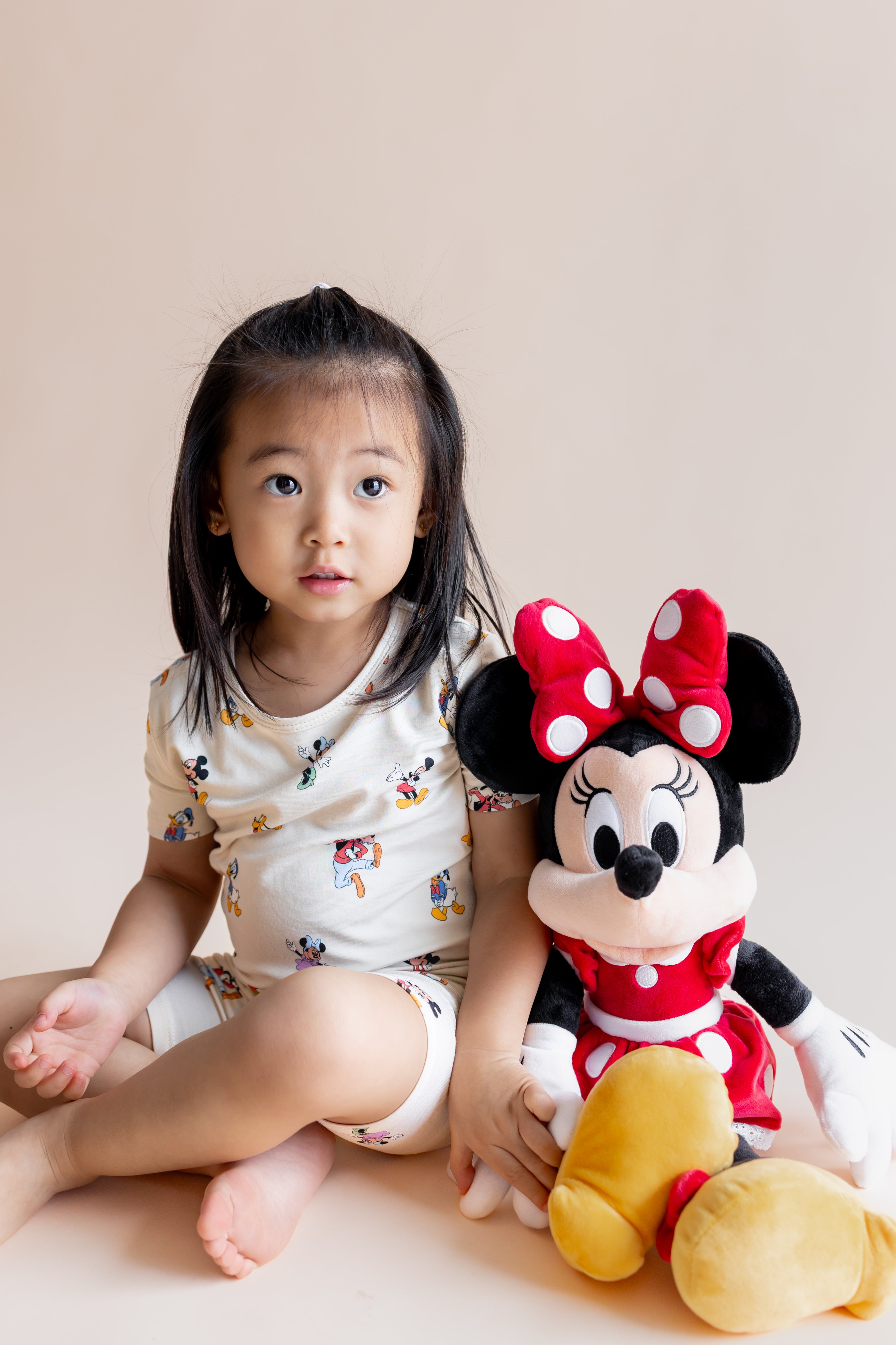 Girl sitting wearing Short Sleeve Pajamas in Mickey Mouse & Friends print with a stuff toy on a cream background
