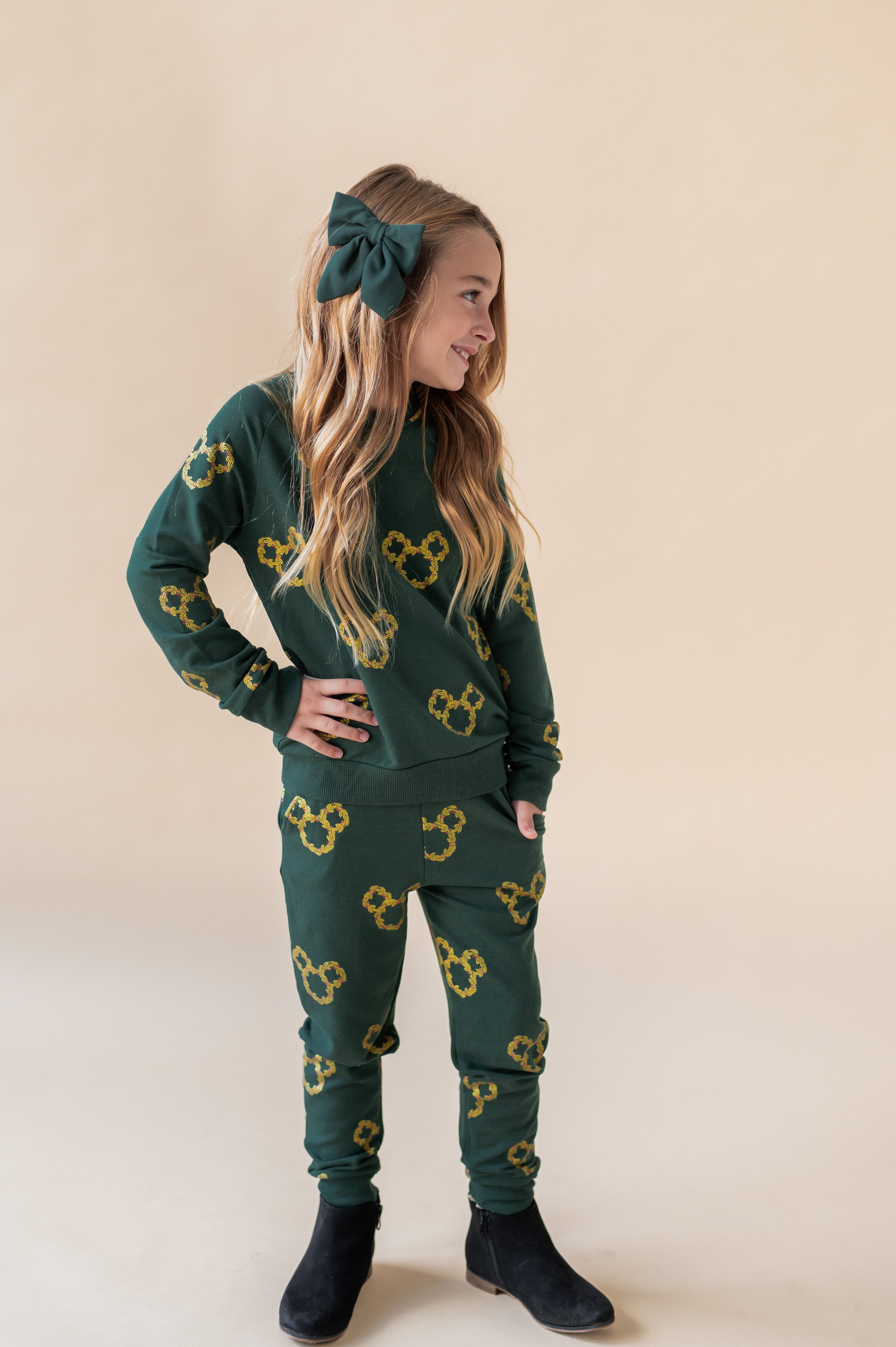Girl looking side ways wearing Long Sleeve Jogger Set in Mickey Christmas Wreath print and green alligator clip bow on a beige background