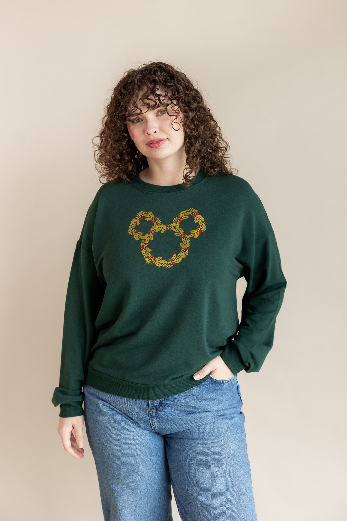 Women wearing Women's Sweatshirt in Mickey Christmas Wreath print on Cypress color on blue jeans on a beige background
