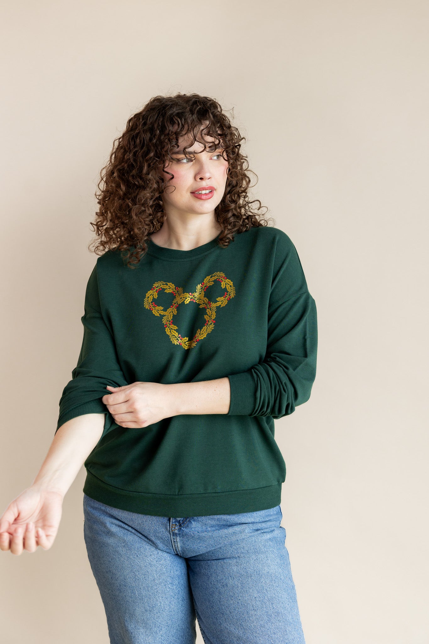Women wearing Women's Sweatshirt in Mickey Christmas Wreath print on Cypress color over blue jeans on a beige background