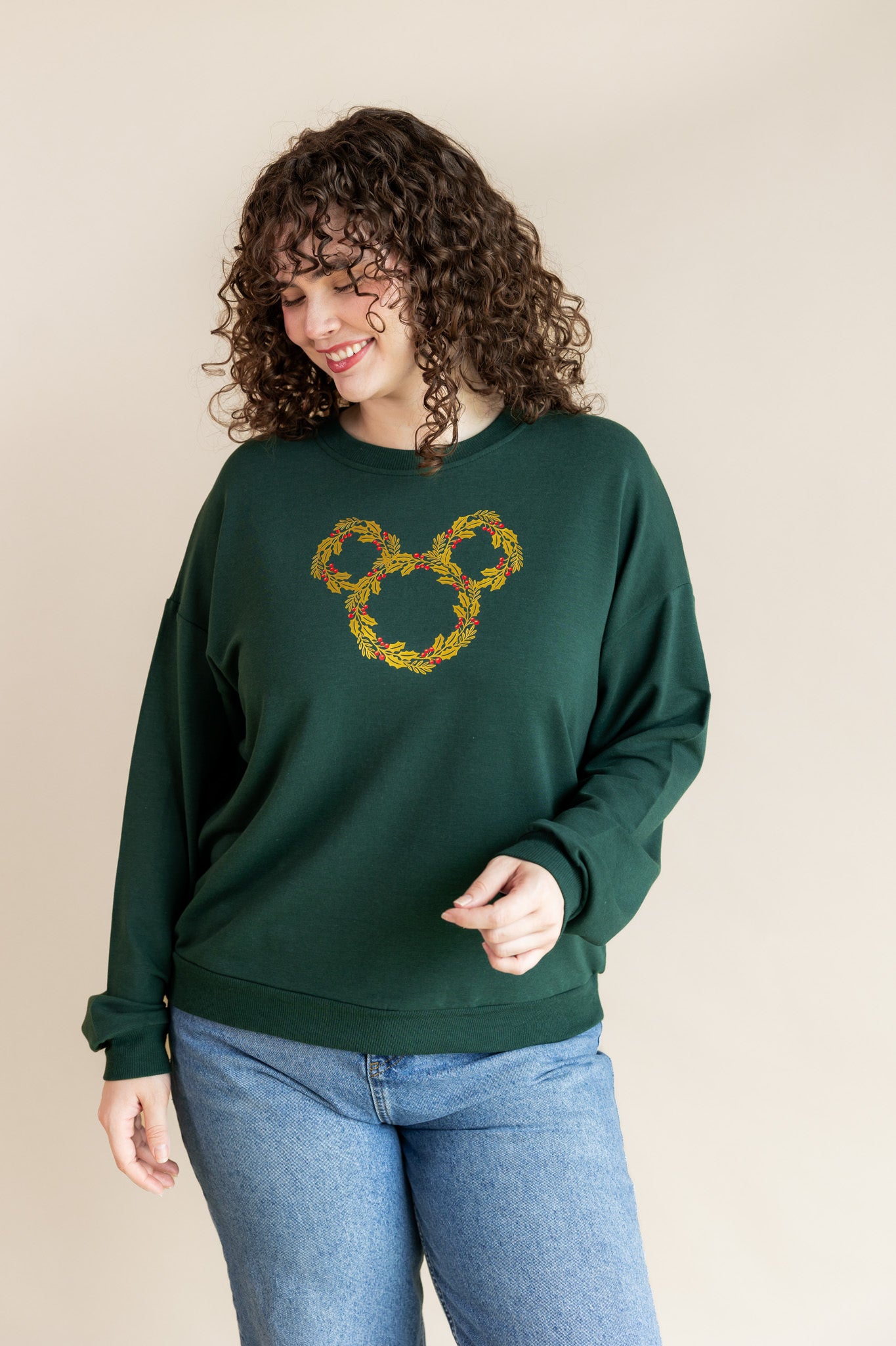 Women smiling in Women's Sweatshirt in Mickey Christmas Wreath print on Cypress color over blue jeans on a beige background