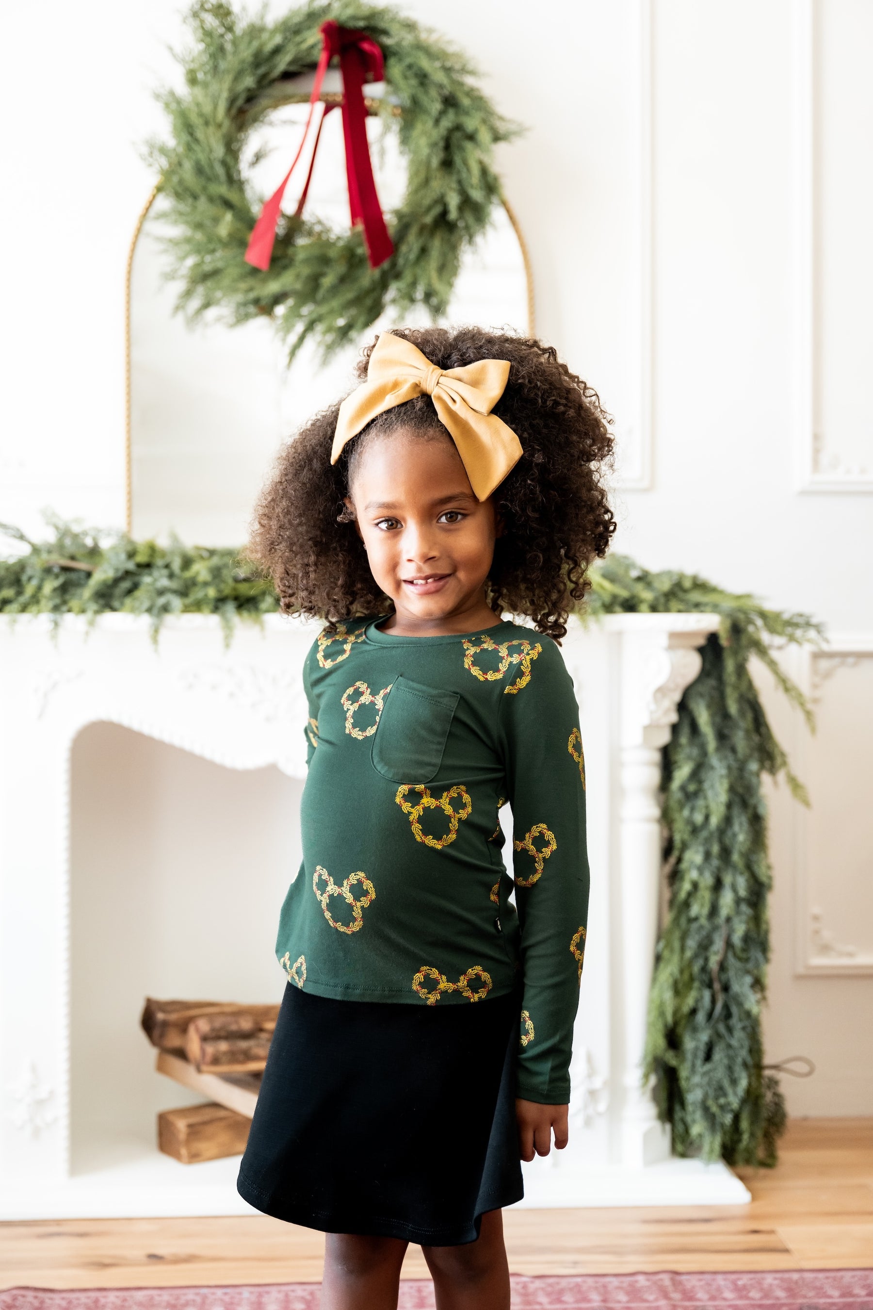 Girl wearing Long Sleeve Toddler Crew Neck Tee in Mickey Christmas Wreath print over black skirt standing in front of fire place