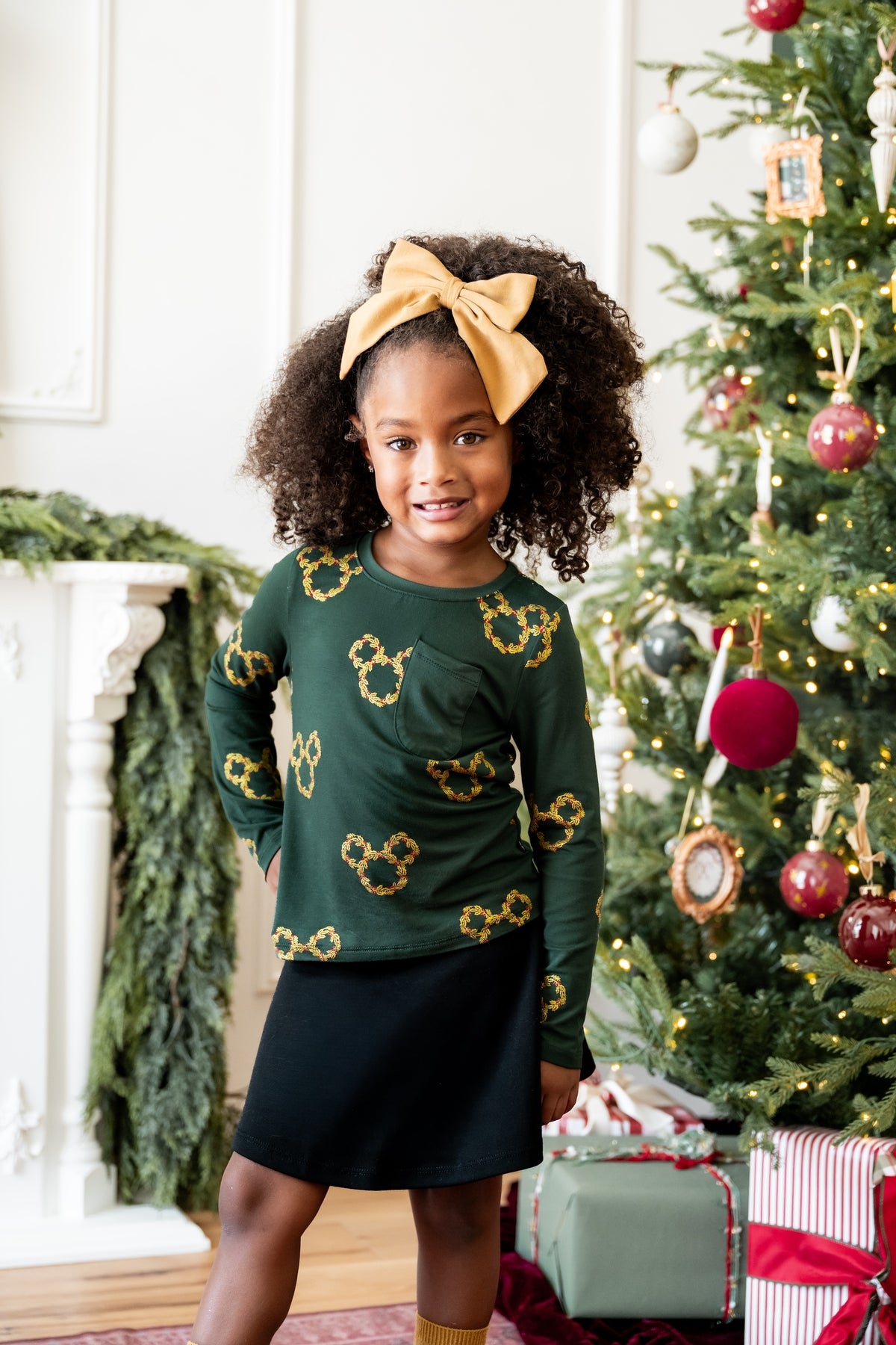 Girl wearing Long Sleeve Toddler Crew Neck Tee in Mickey Christmas Wreath print over black skirt standing beside the Chriistmas tree