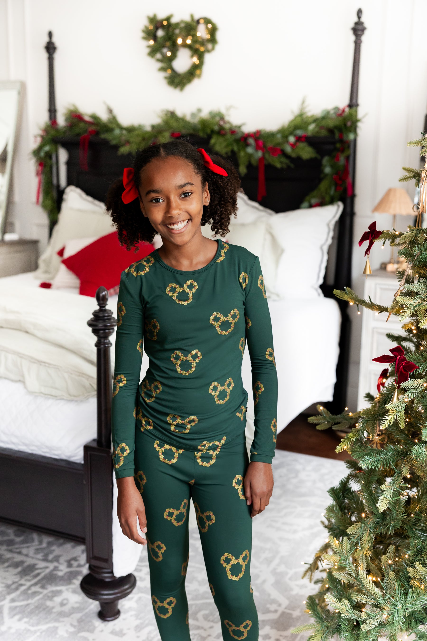 A young girl smiling in a green long-sleeve pajama set with gold Mickey Mouse head patterns, standing in a decorated bedroom next to a Christmas tree.