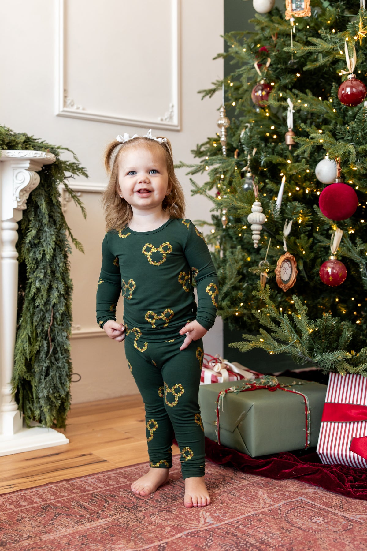 A young girl stands barefoot in a living room next to a decorated Christmas tree, wearing a dark green long-sleeve pajama set with gold Mickey Mouse head patterns.