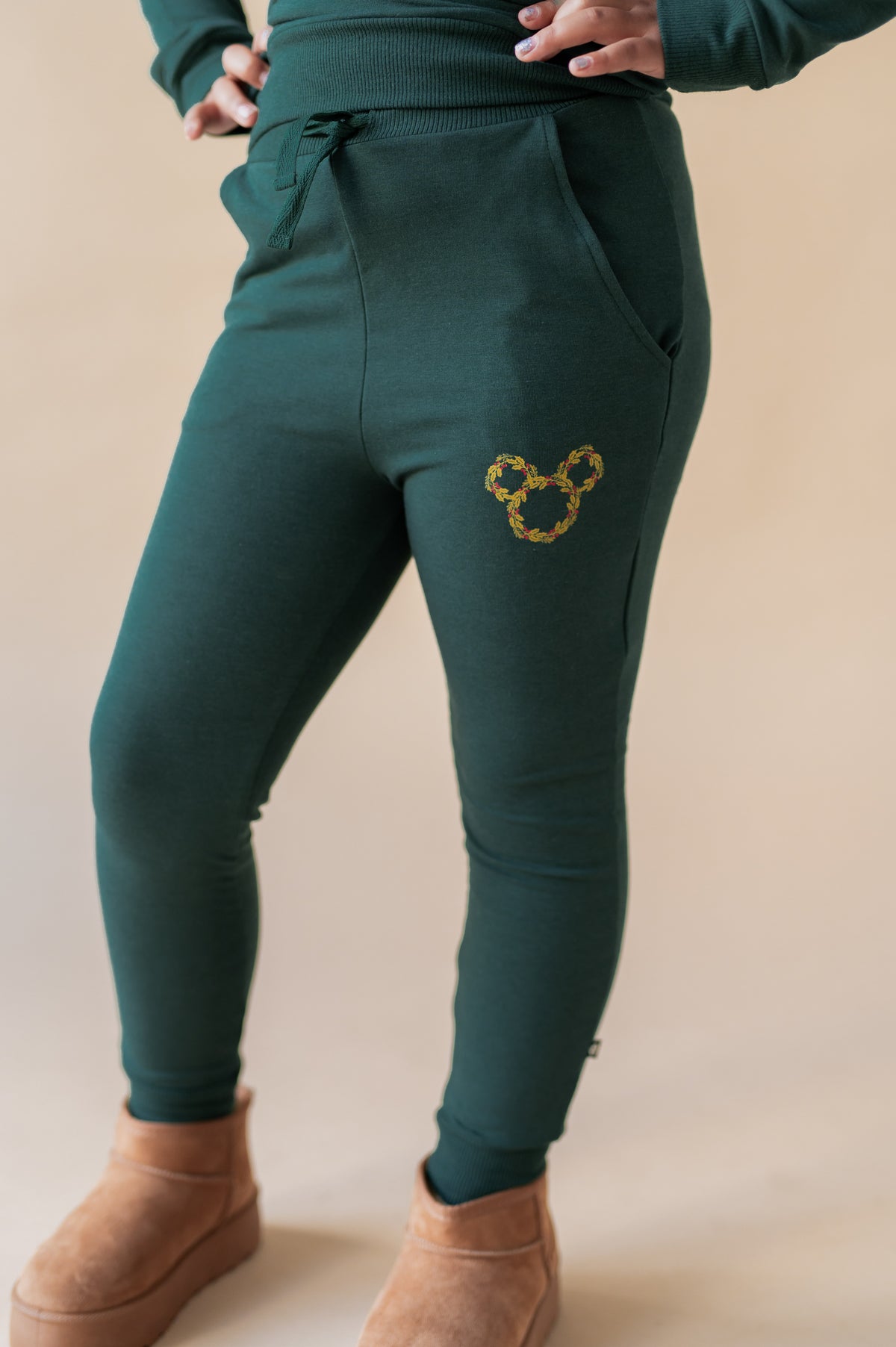 Girl showcasing pants of Long Sleeve Jogger Set with Mickey Christmas Wreath print on the side in cypress color on a beige background