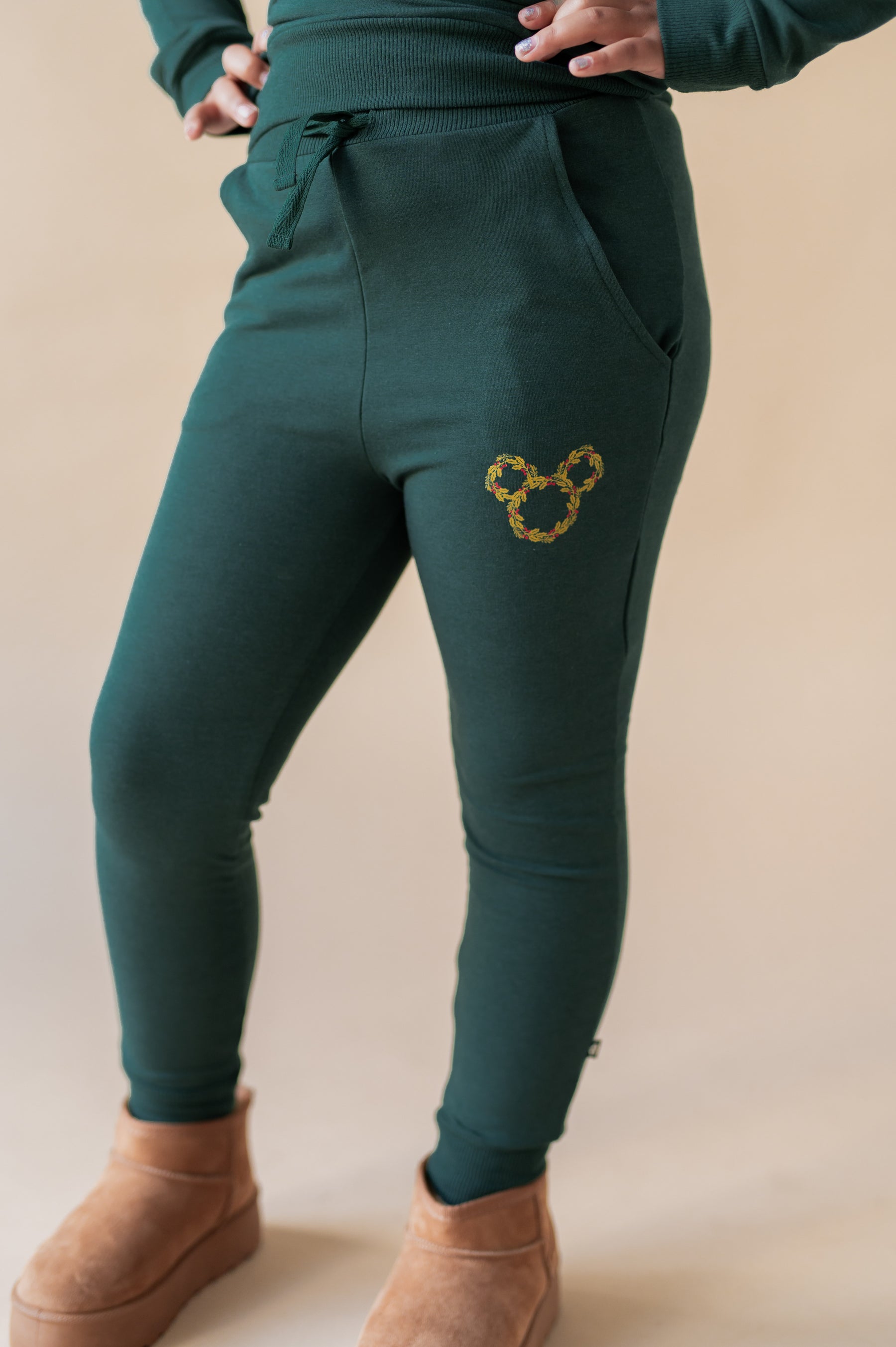 Girl showcasing pants of Long Sleeve Jogger Set with Mickey Christmas Wreath print on the side in cypress color on a beige background