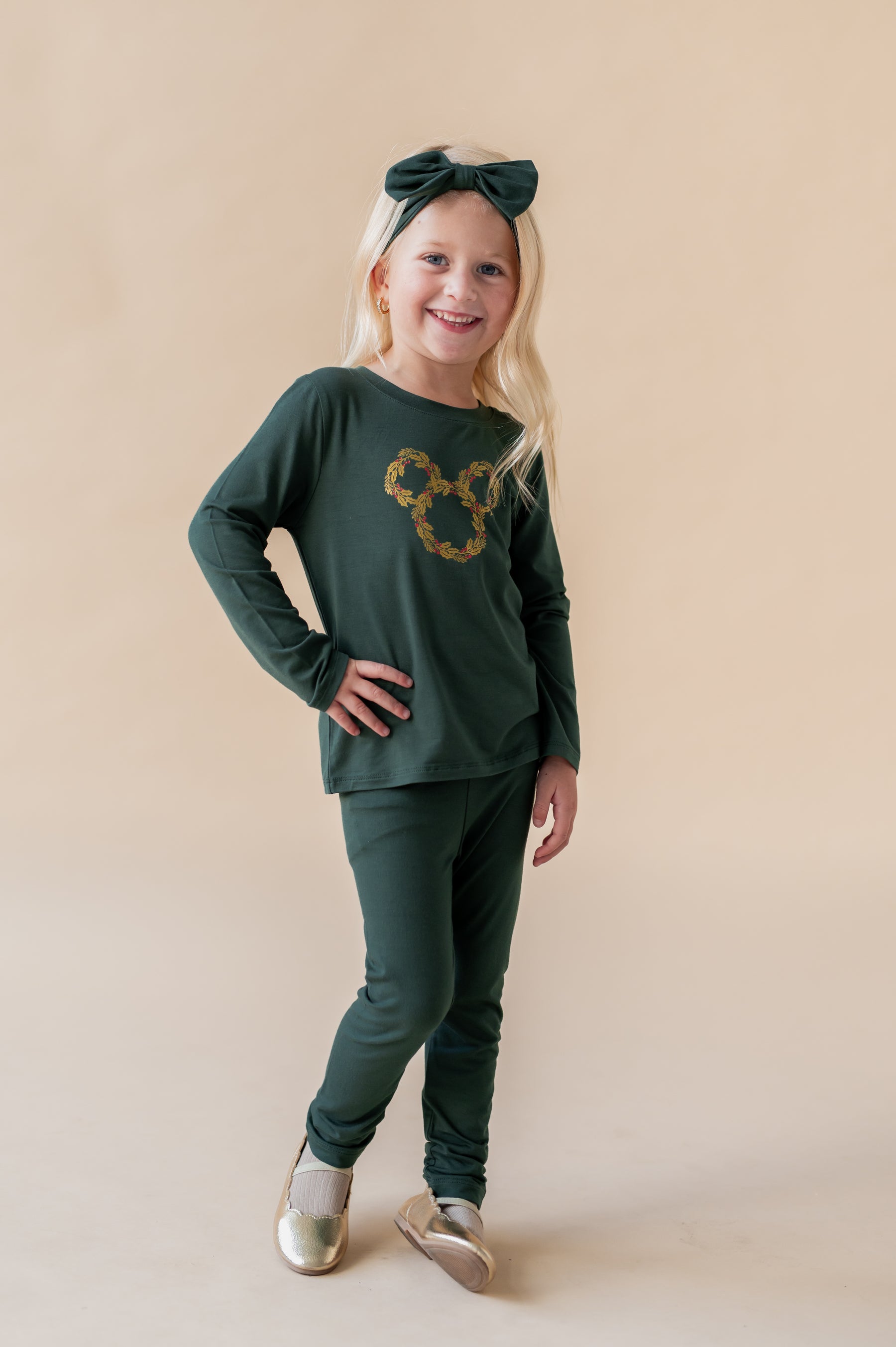 Girl posing in Long Sleeve Toddler Crew Neck Tee in Mickey Christmas Wreath print in center on Cypress color tee over similar color leggings and knotted bow headband on a beige background