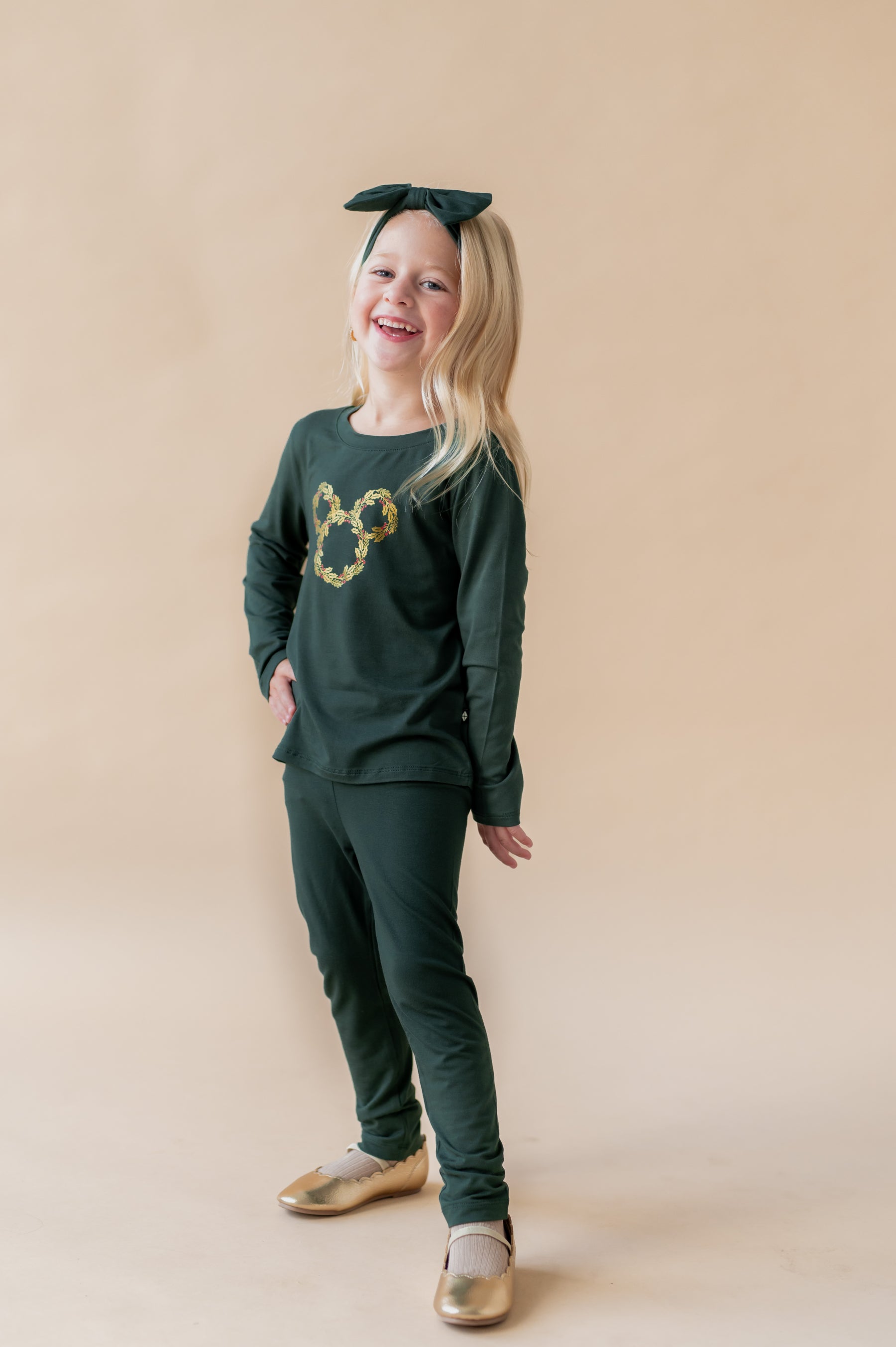 Girl laughing while wearing Long Sleeve Toddler Crew Neck Tee in Mickey Christmas Wreath print in center on Cypress color tee over similar color leggings and knotted bow headband on a beige background