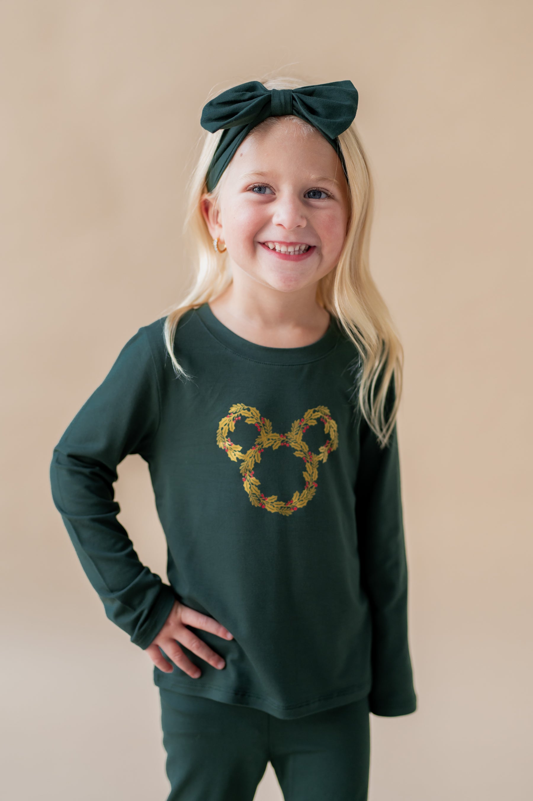 Girl wearing Long Sleeve Toddler Crew Neck Tee in Mickey Christmas Wreath print in center on Cypress color tee over similar color leggings and knotted bow headband on a beige background