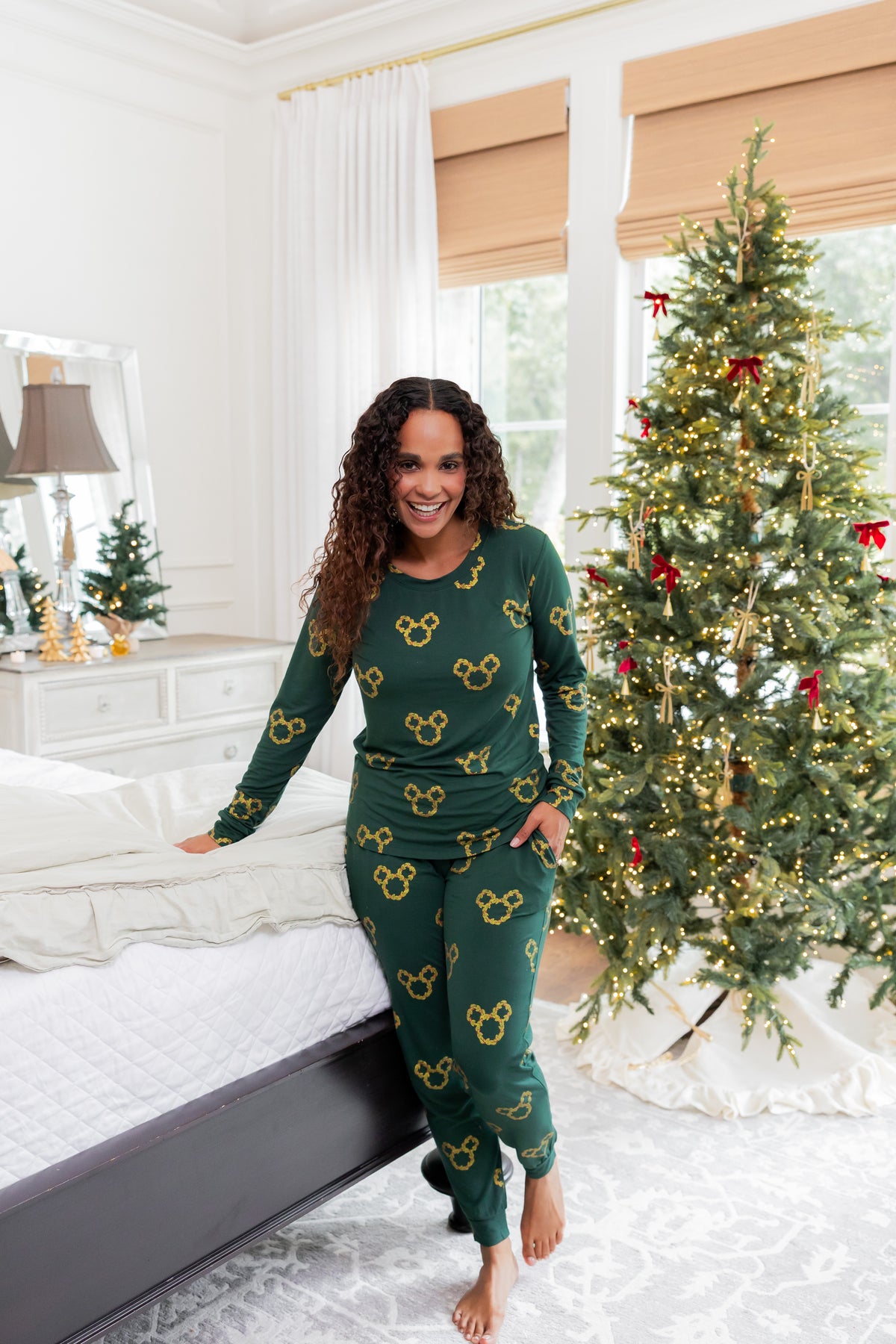 Woman wearing Women's Jogger Pajama Set in Mickey Christmas Wreath print smiling in front of Christmas tree in a bedroom