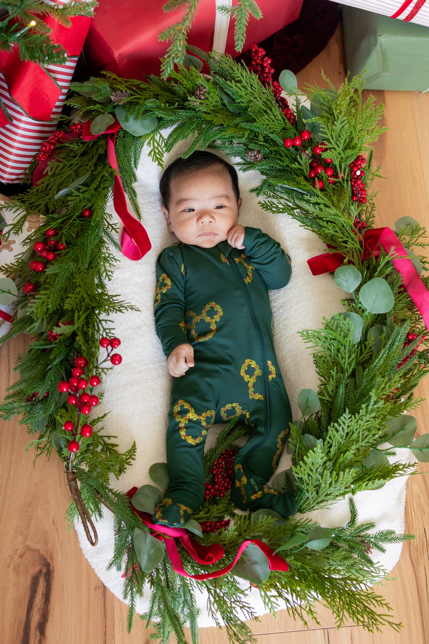 Baby wearing Zippered Footie in Mickey Christmas Wreath print lying between presents decoration