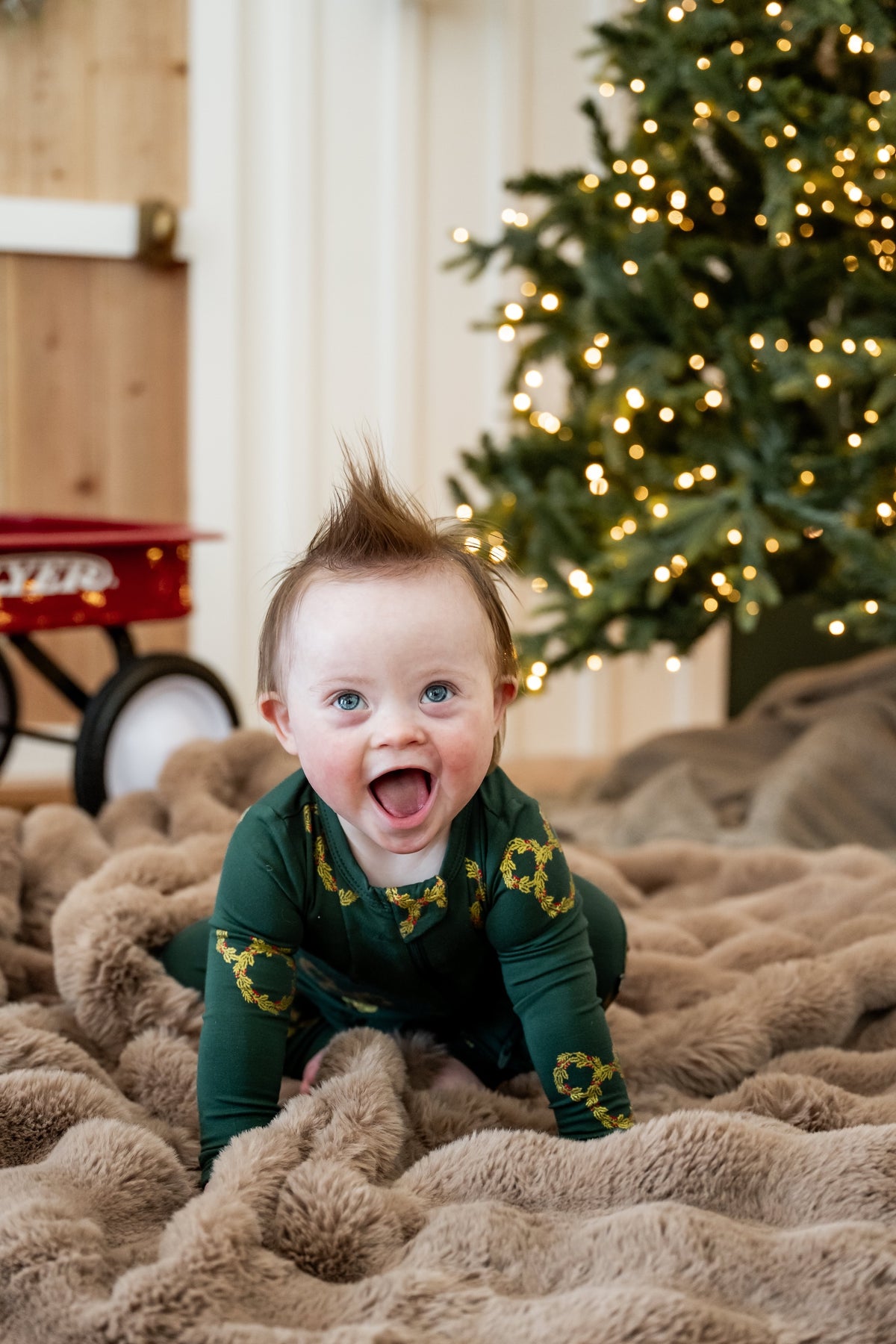 Baby wearing Zippered Romper in Mickey Christmas Wreath print sitting on a beige blanket in front of a Christmas tree