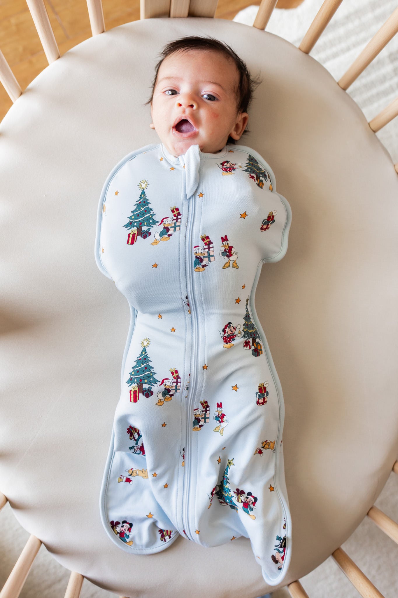 Baby lying in crib wearing Swaddle Bag in Mickey & Friends Christmas 0.5 print on beige crib sheet