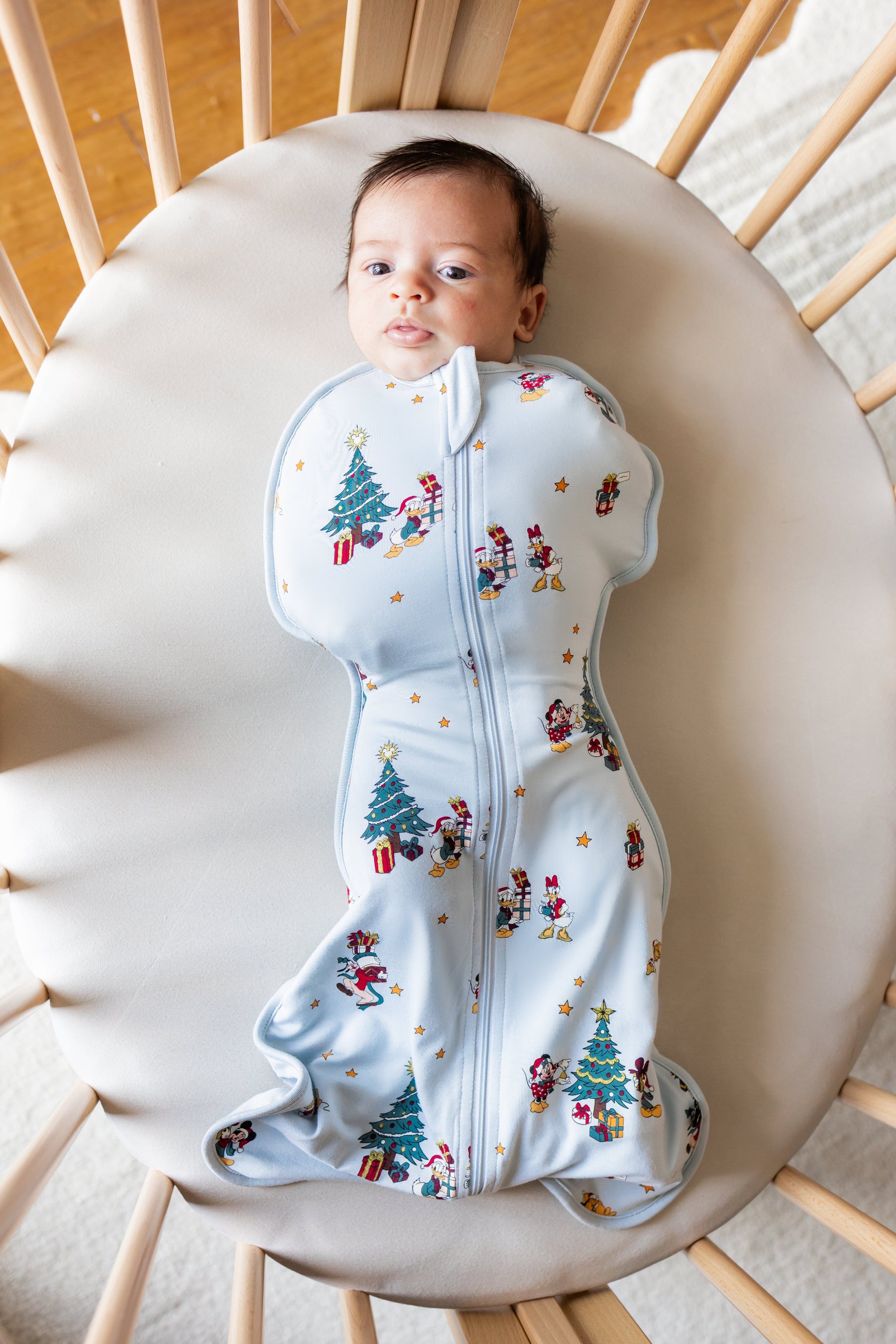 Baby lying in crib wearing Swaddle Bag in Mickey & Friends Christmas 0.5 print over beige crib sheet