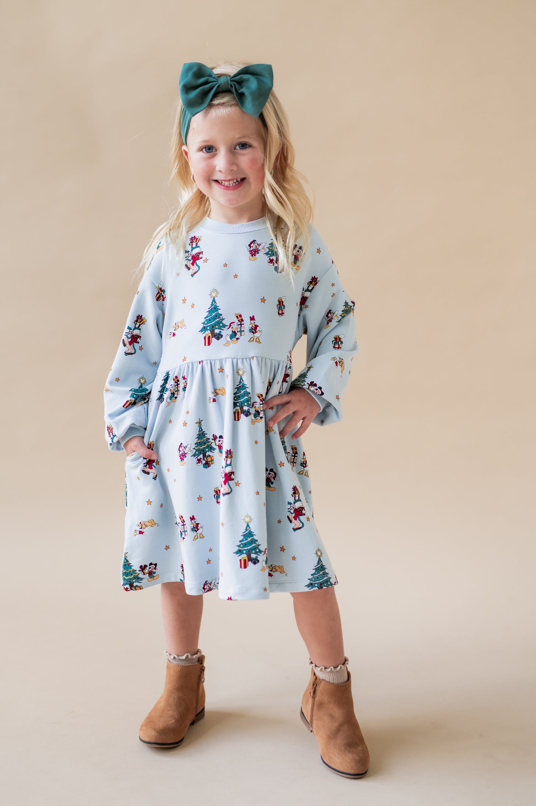 Girl wearing Long Sleeve Skater Dress Mickey & Friends Christmas print with green knotted bow headband on brown boots on a beige background