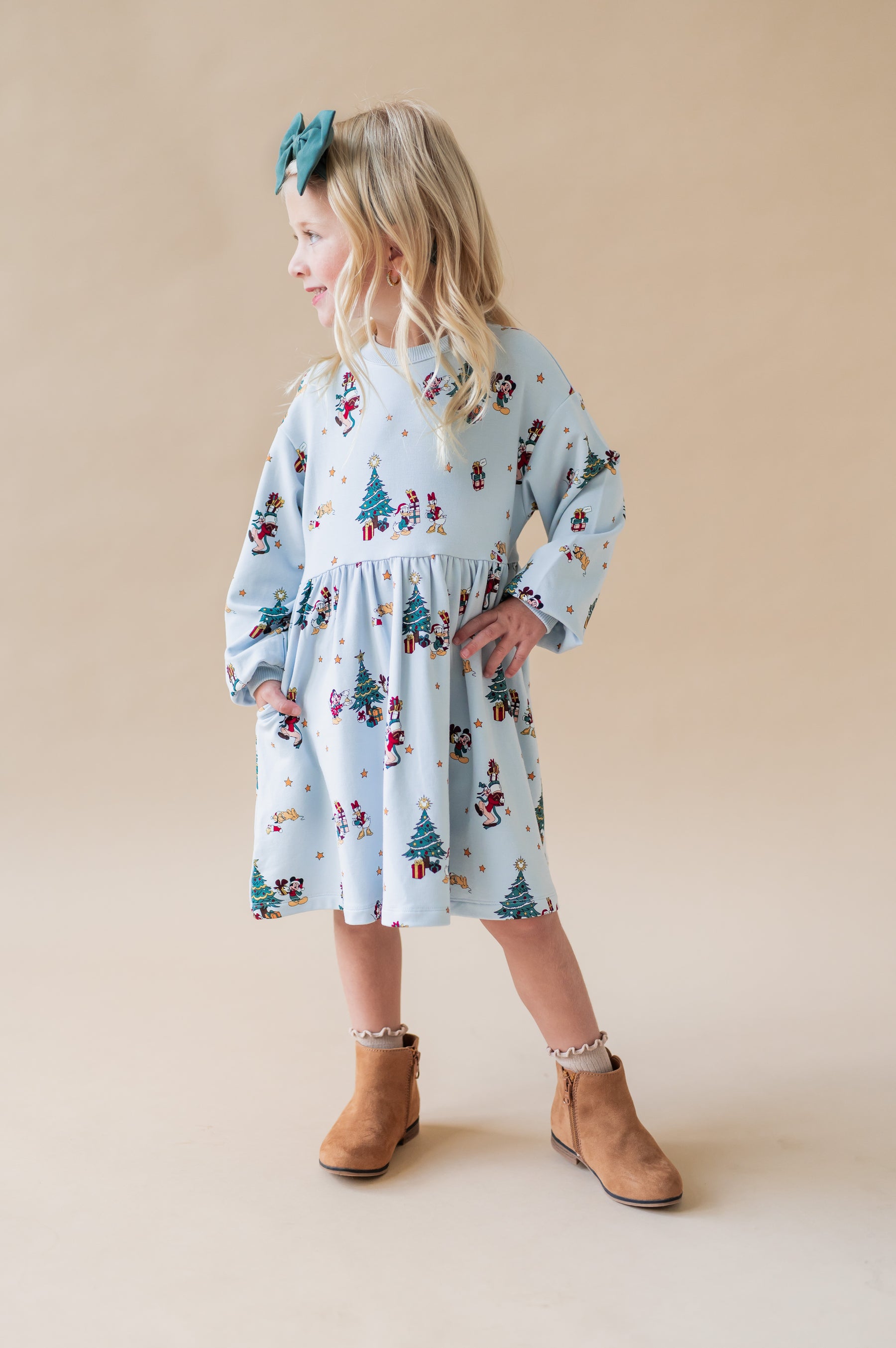 Girl facing side ways wearing Long Sleeve Skater Dress Mickey & Friends Christmas print and green bow on brown boots on a beige background