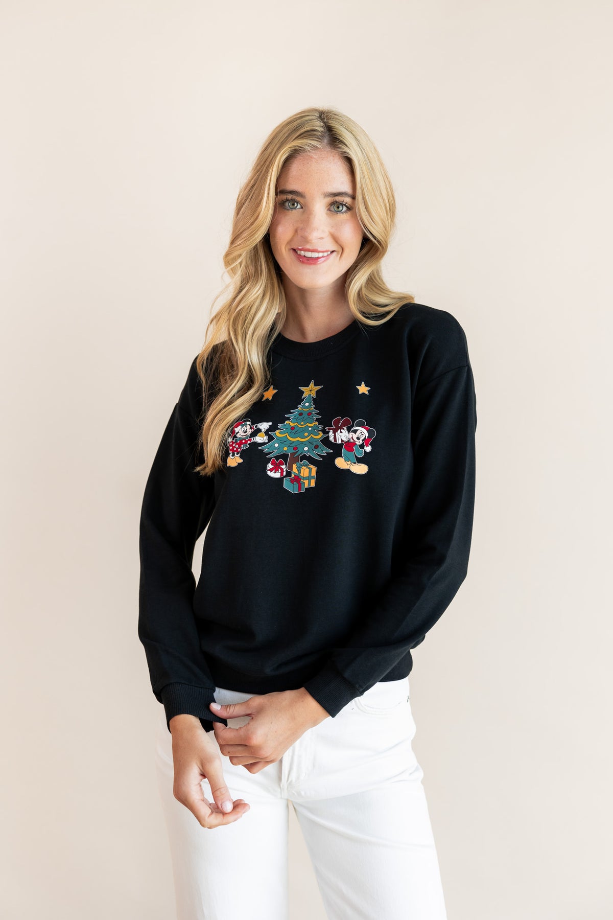 Woman wearing Black Women's Sweatshirt in Mickey & Friends Christmas print in the center over white pants on a beige background