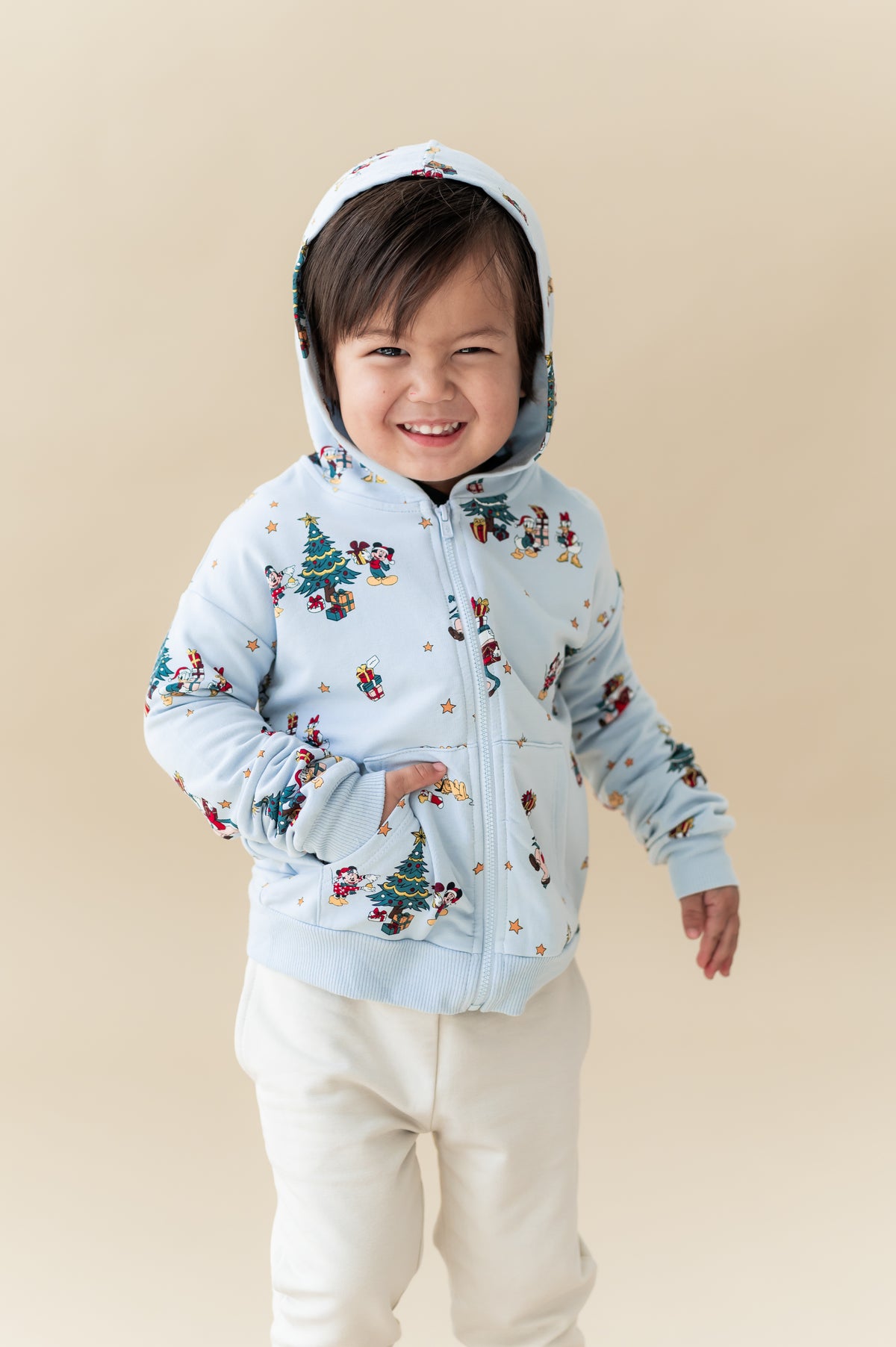 Boy wearing Zip-Up Hoodie in Mickey & Friends Christmas print over white pants on a beige background