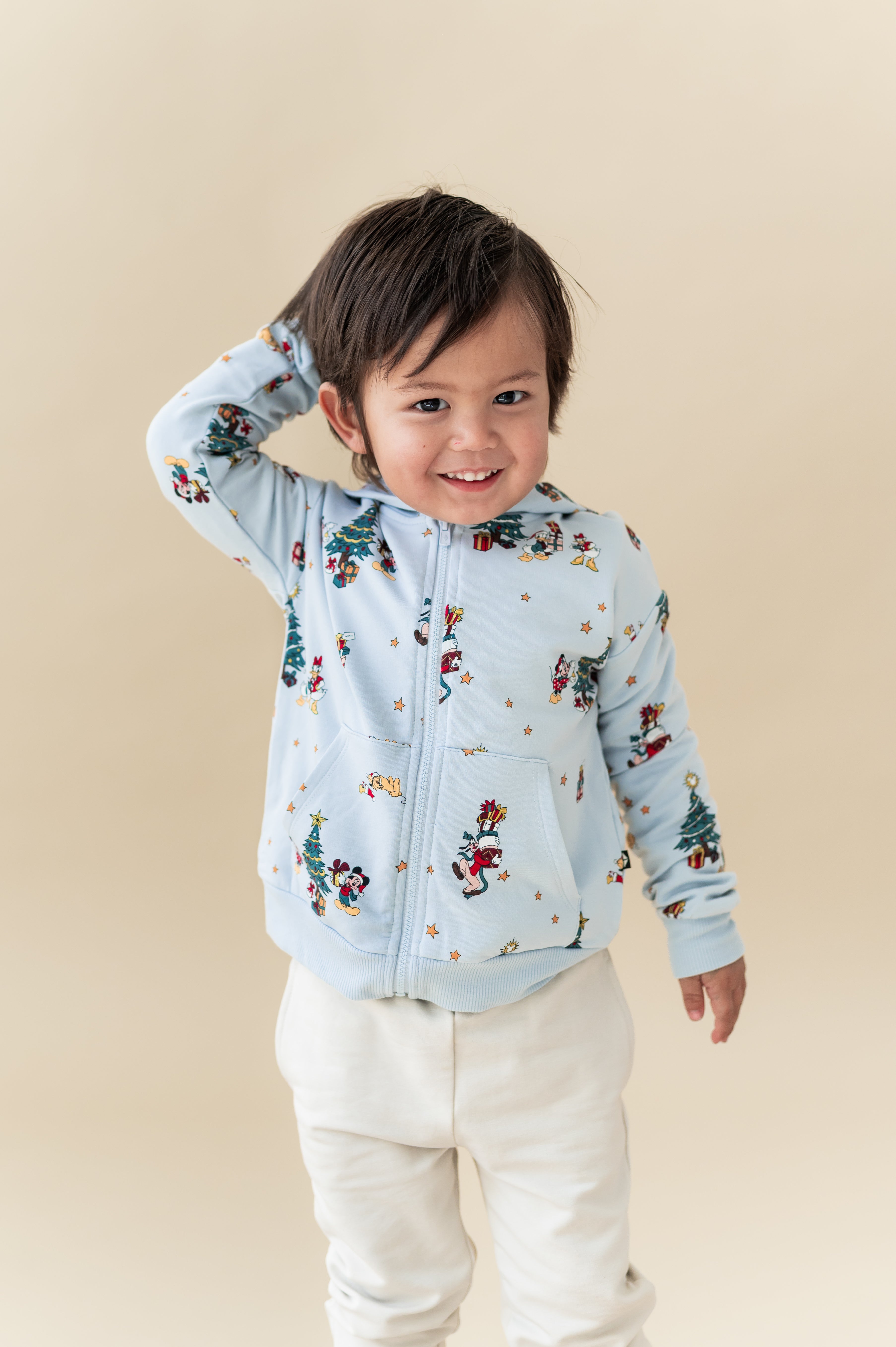 Boy wearing Zip-Up Hoodie in Mickey & Friends Christmas print over white pants on a beige background