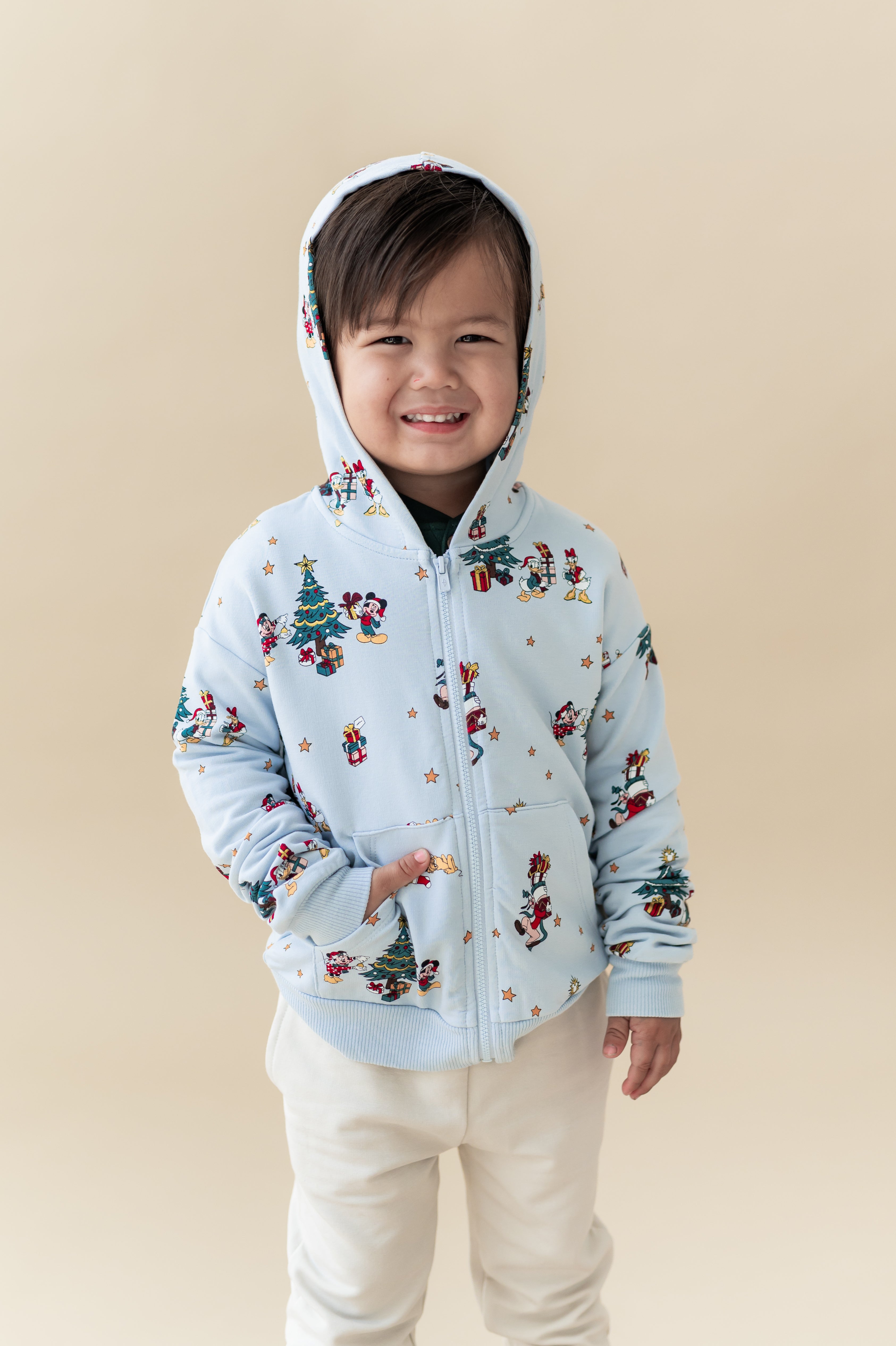 Boy smiling in Zip-Up Hoodie in Mickey & Friends Christmas print over white pants on a beige background