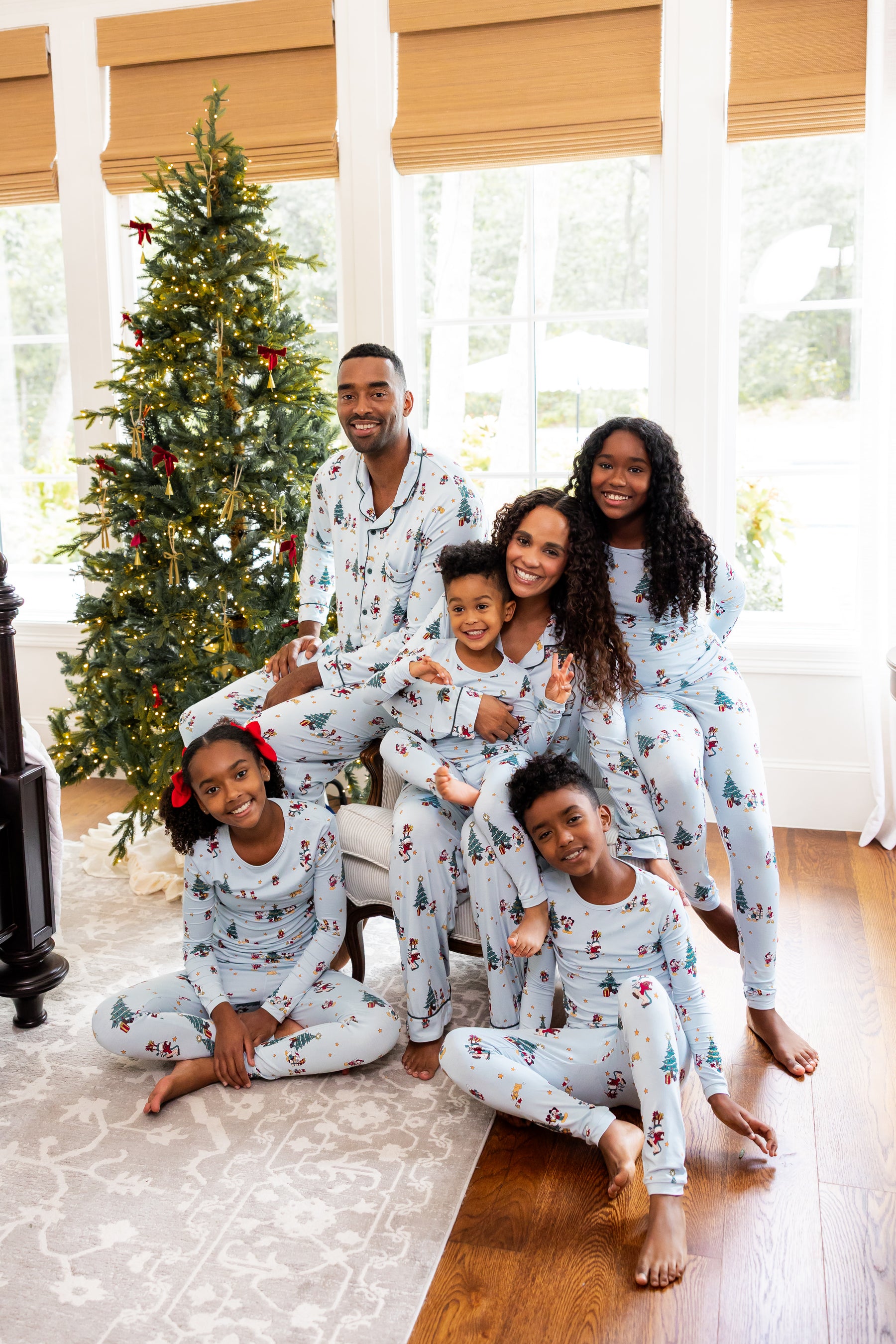 A family of six wearing matching light blue Christmas-themed disney pajamas poses together in a bright living room with a decorated Christmas tree, large windows, and wood floors.